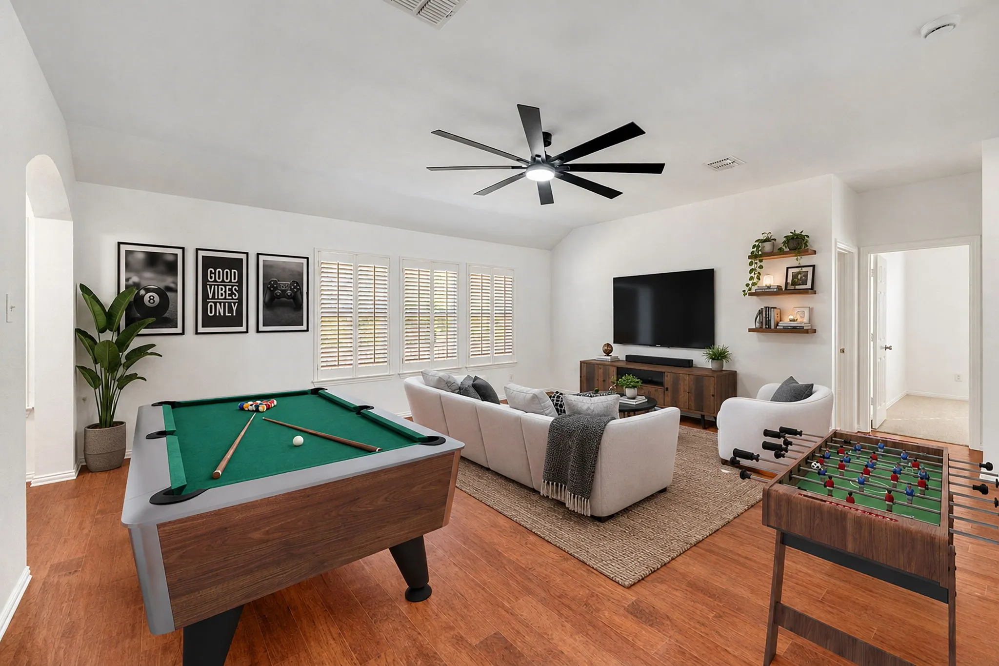 Spacious living area featuring wood-finish flooring, multiple windows with plantation shutters, a ceiling fan, and built-in floating shelves
