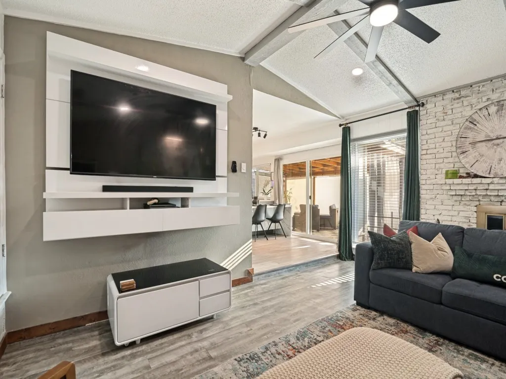 Vaulted ceiling with exposed beams, wood-finish flooring, white brick-style accent wall, built-in media console, and a contemporary ceiling fan with integrated lighting