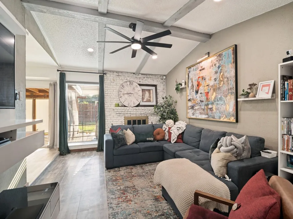 Vaulted ceiling with exposed beams, wood-finish flooring, white brick fireplace, built-in shelving, and recessed lighting