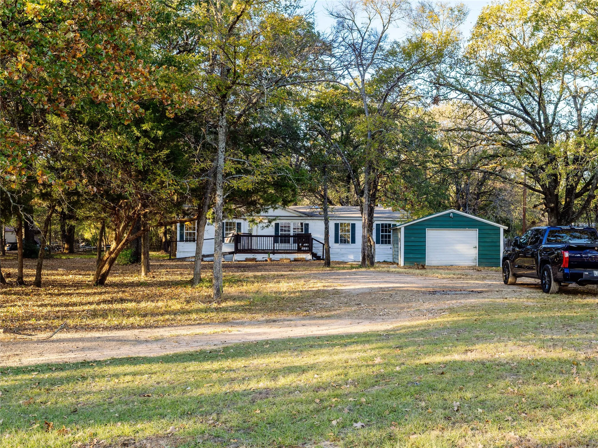 White exterior with blue shutters, front porch with black railing, detached teal garage, gravel driveway, and mature trees