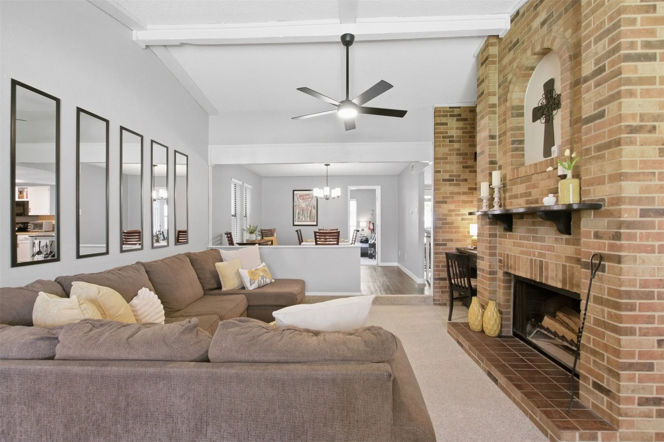 Exposed brick fireplace with an arched niche, vaulted ceiling with exposed beams, ceiling fan, and light-colored carpeting