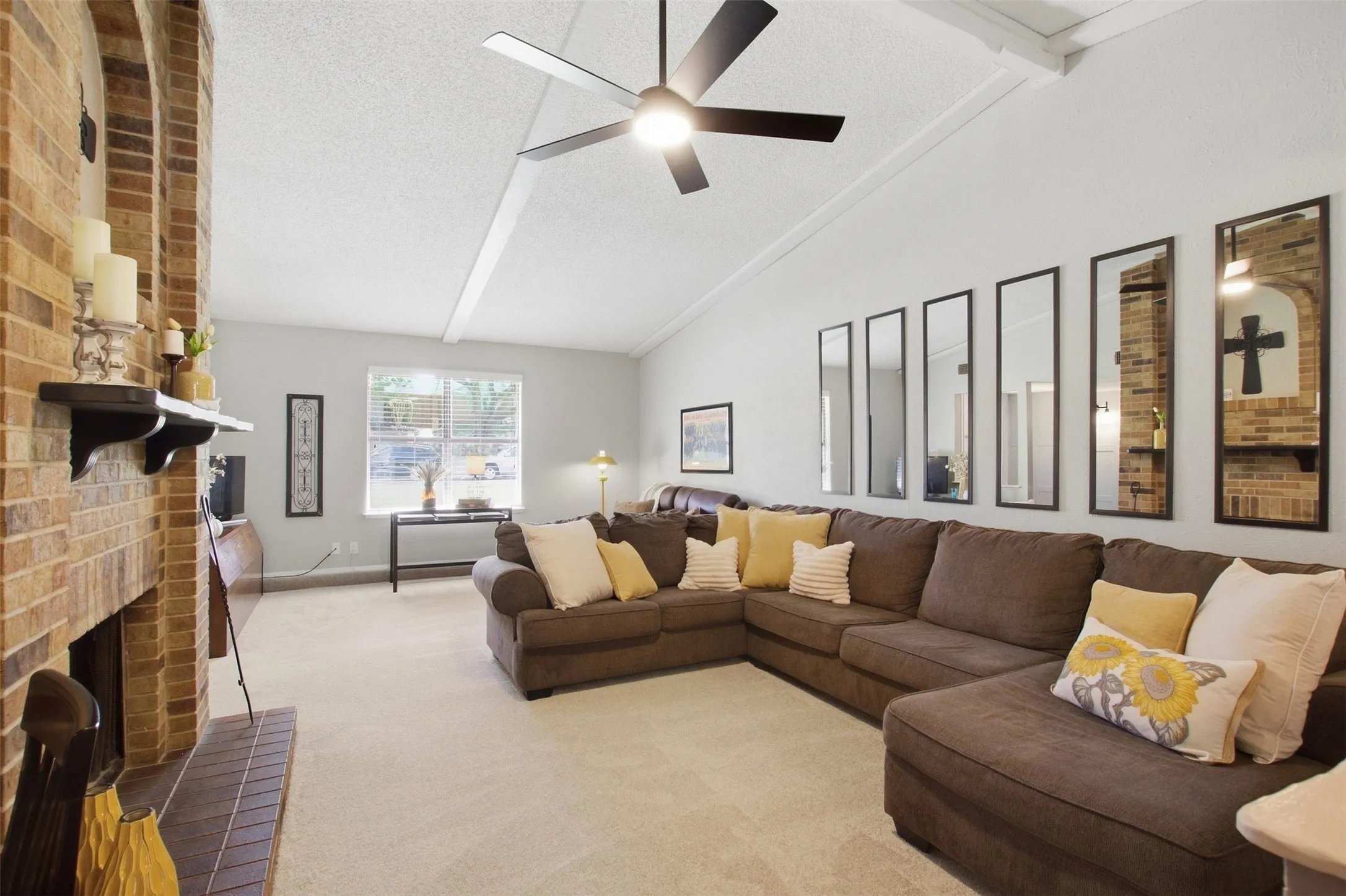 Vaulted ceiling with exposed beams, brick fireplace, ceiling fan with integrated lighting, carpeted flooring, and a large window