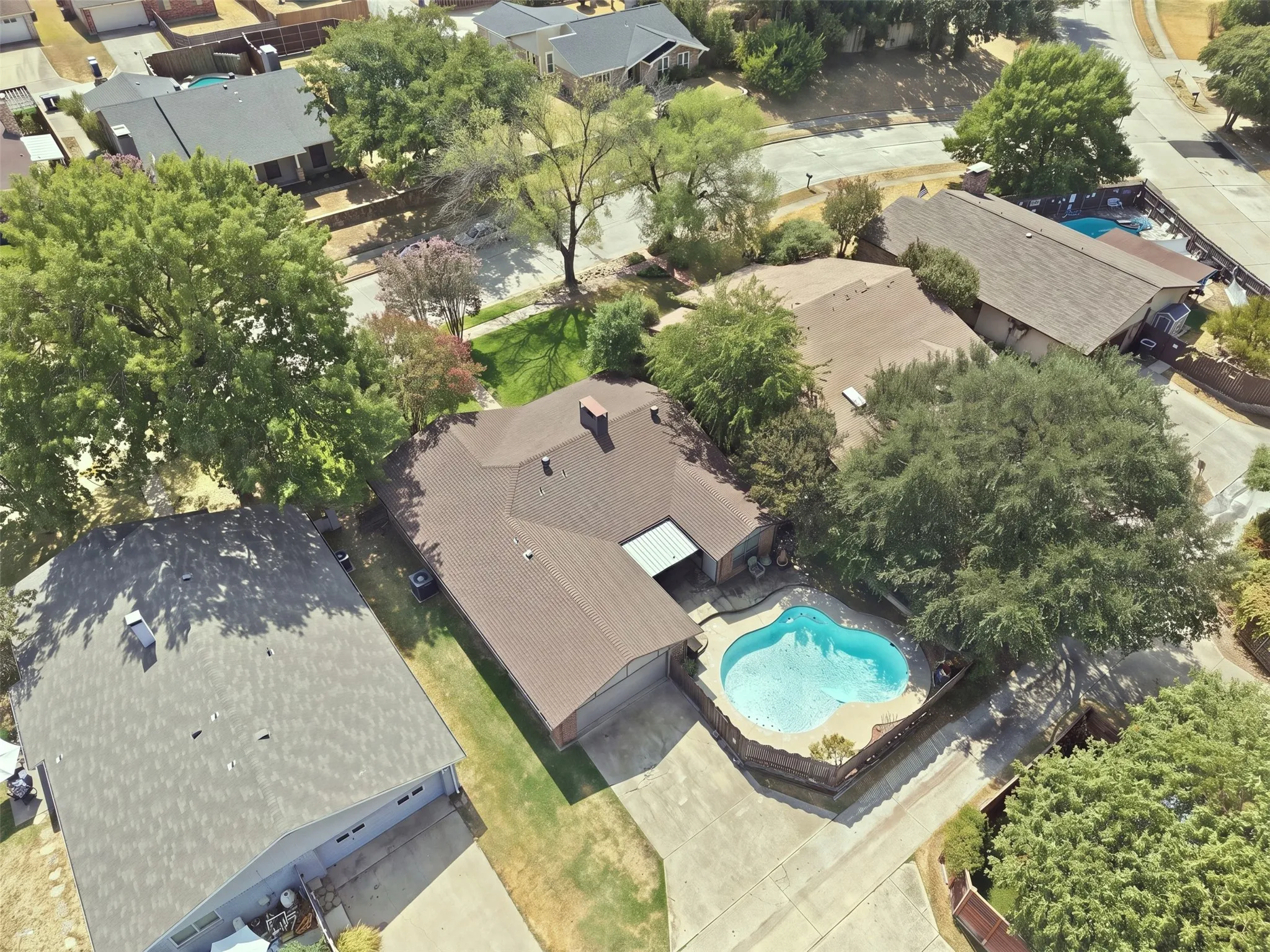 Shingle roof residence with an irregular shape, featuring an in-ground swimming pool, mature trees, and concrete driveways