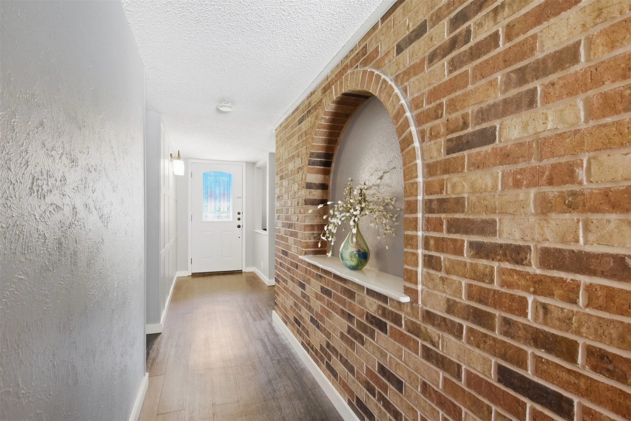 Brick accent wall with an arched niche, wood-finish flooring, textured wall finish, and a white paneled door with an arched window