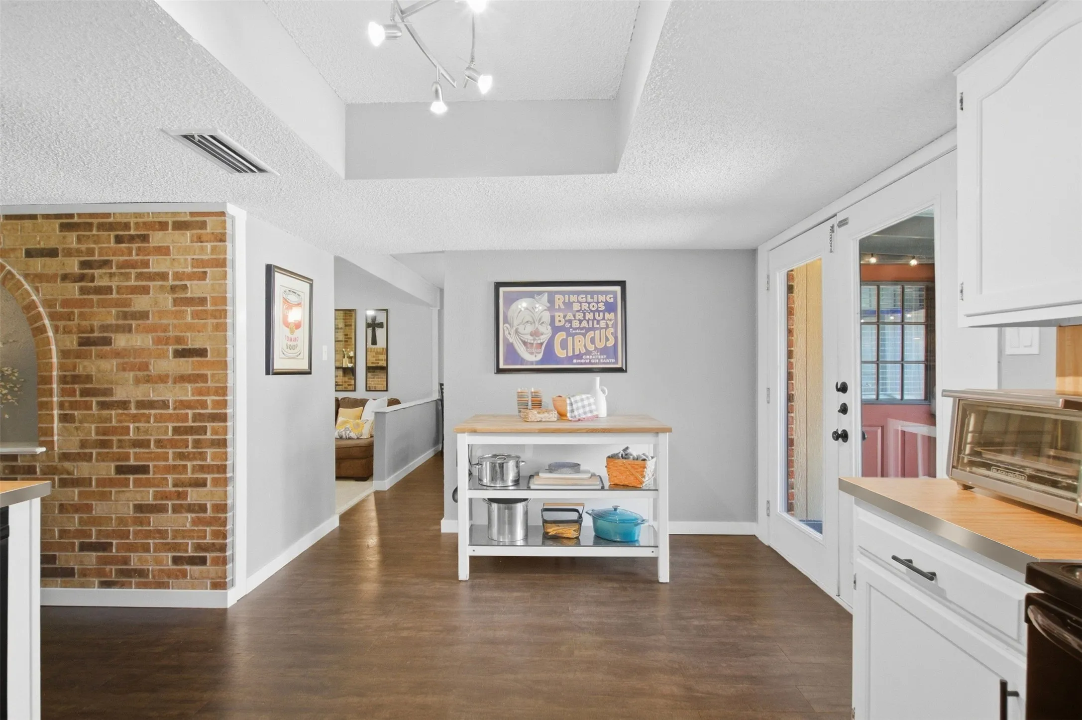 Wood-finish flooring throughout, brick accent wall with arched opening, white cabinetry with dark hardware, recessed ceiling feature, and track lighting
