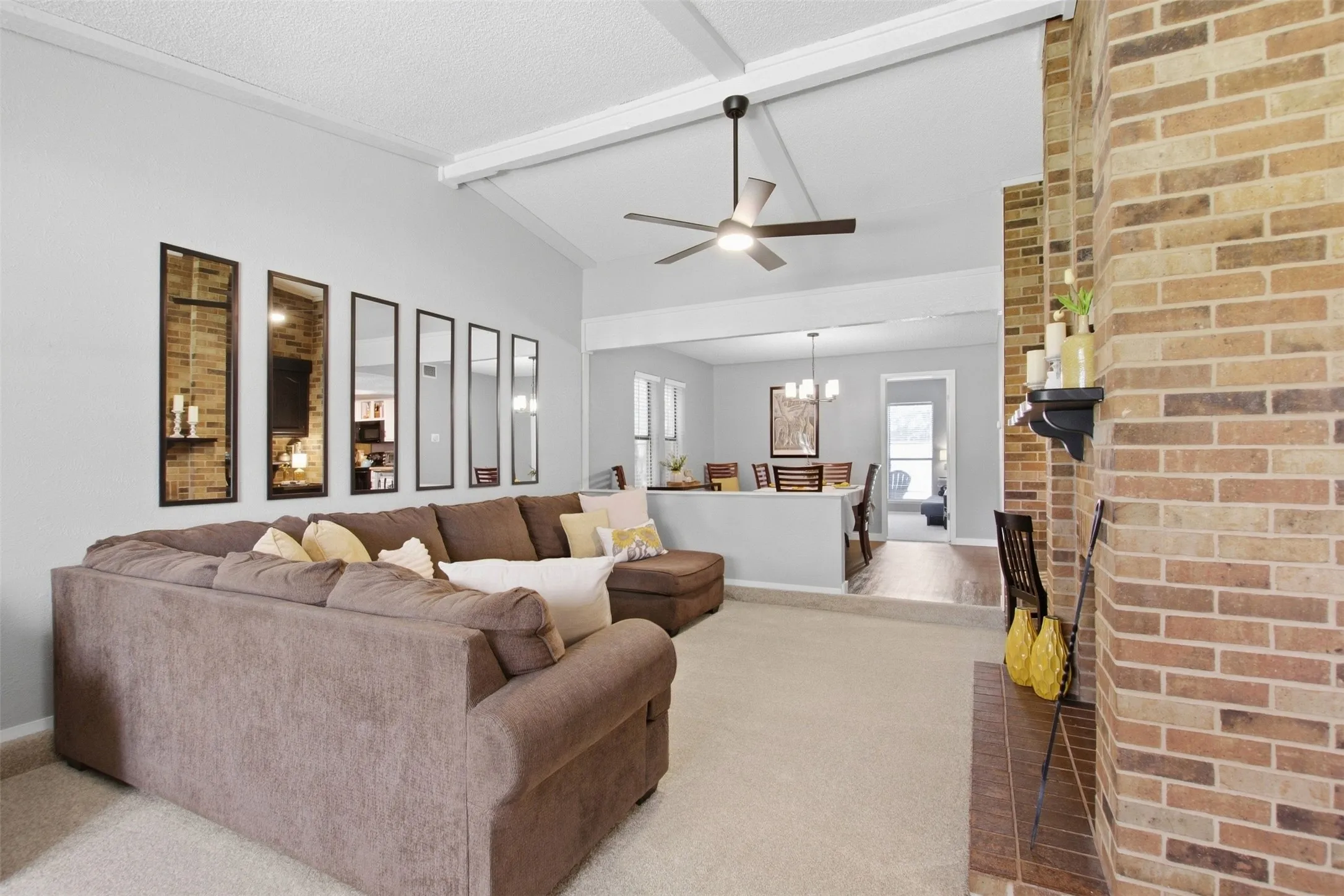 Vaulted ceiling with exposed beams, brick-faced fireplace, ceiling fan with integrated lighting, carpeted flooring, and a partial wall separating spaces
