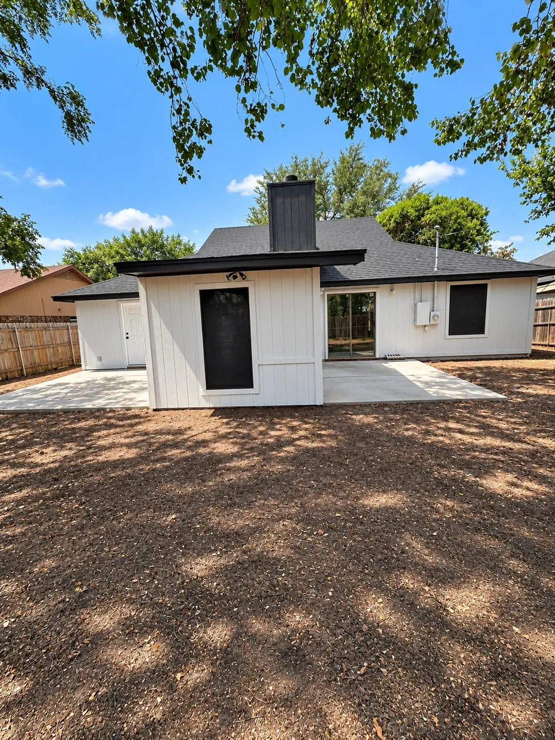 Exterior featuring white vertical siding, black trim, a dark asphalt shingle roof, a prominent dark chimney, and multiple concrete patio areas