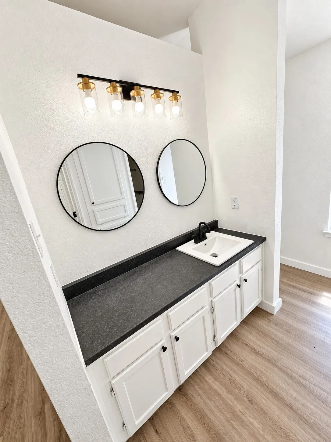 Wood-finish flooring, white vanity cabinetry with black hardware, dark countertop, white sink, and a black faucet