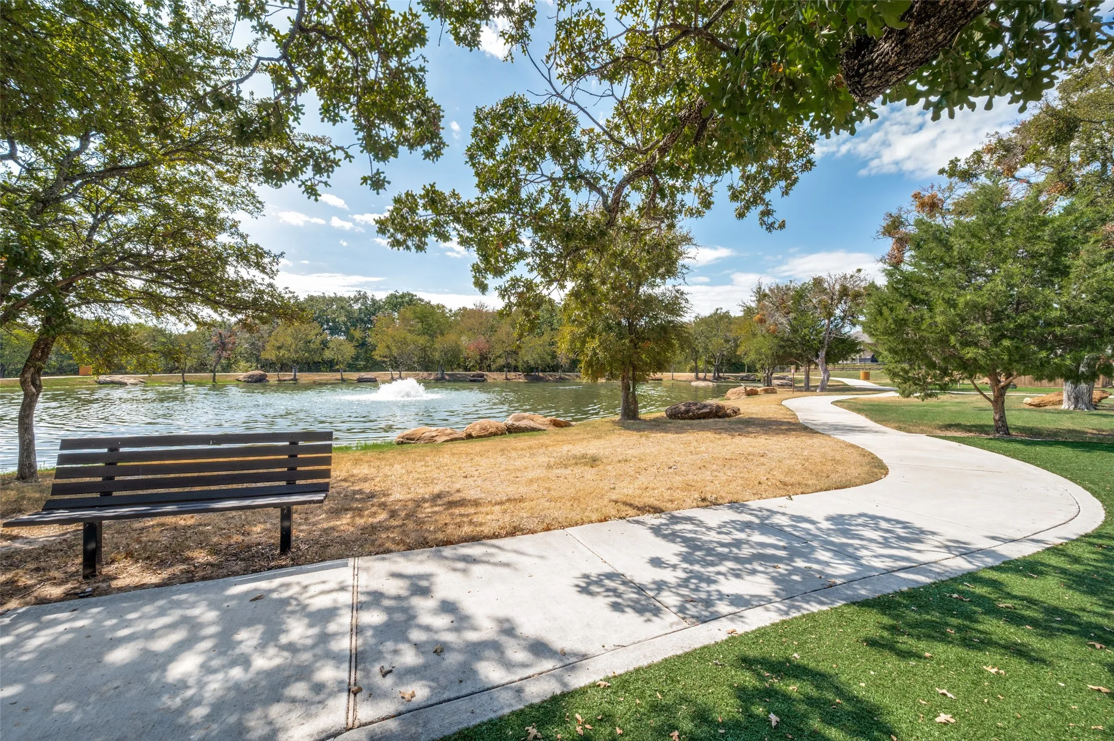 Curved concrete pathway, mature trees, expansive green space, tranquil pond with fountain, and a permanent bench