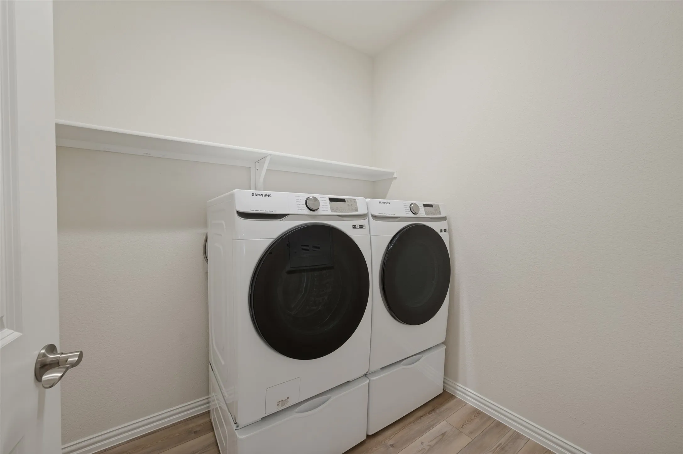 Dedicated laundry area featuring a white storage shelf, light-toned wood-finish flooring, and a contemporary door with a brushed nickel handle