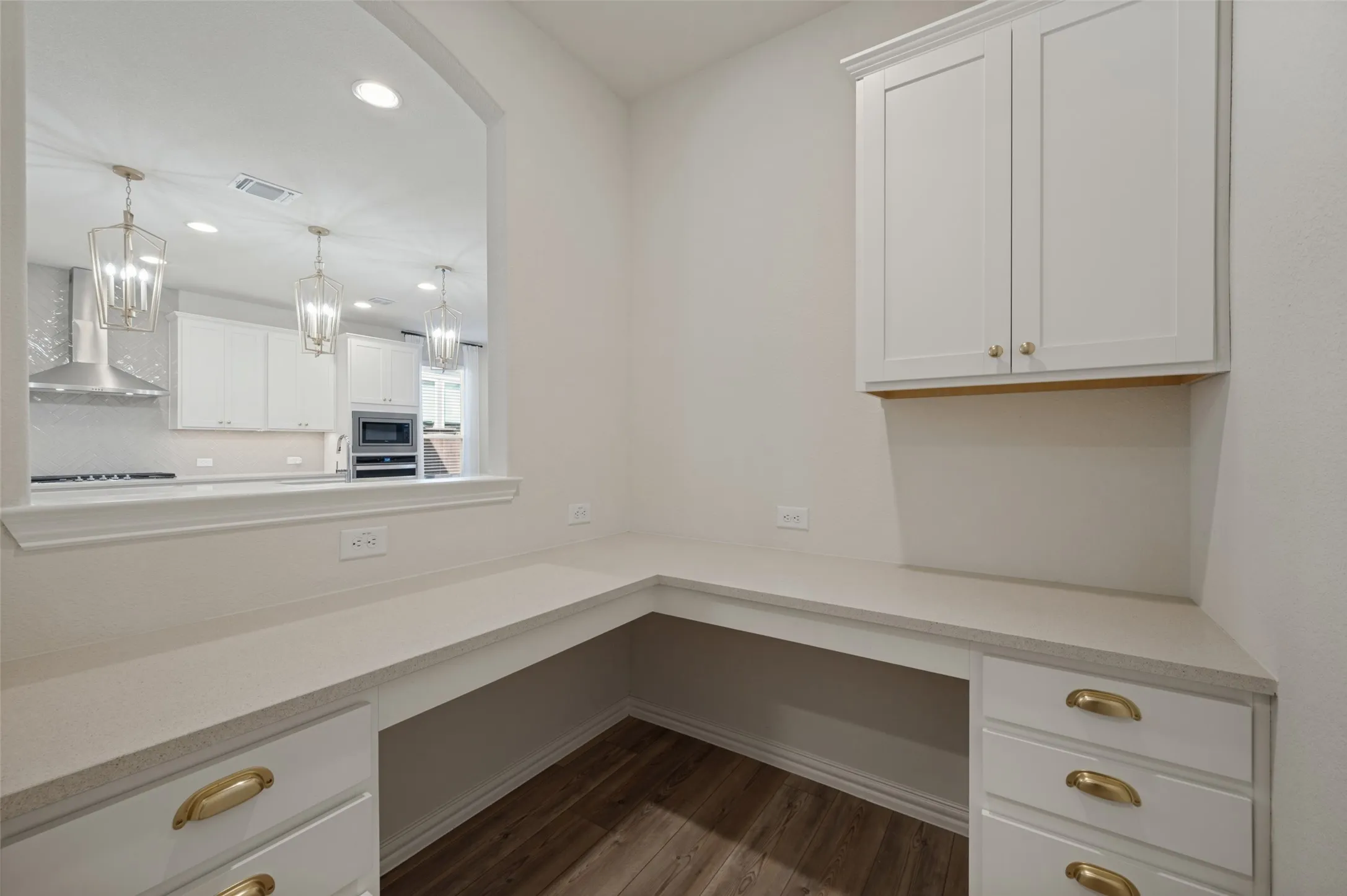 Built-in desk with overhead cabinetry, L-shaped countertop, white cabinetry with gold hardware, and wood-finish flooring