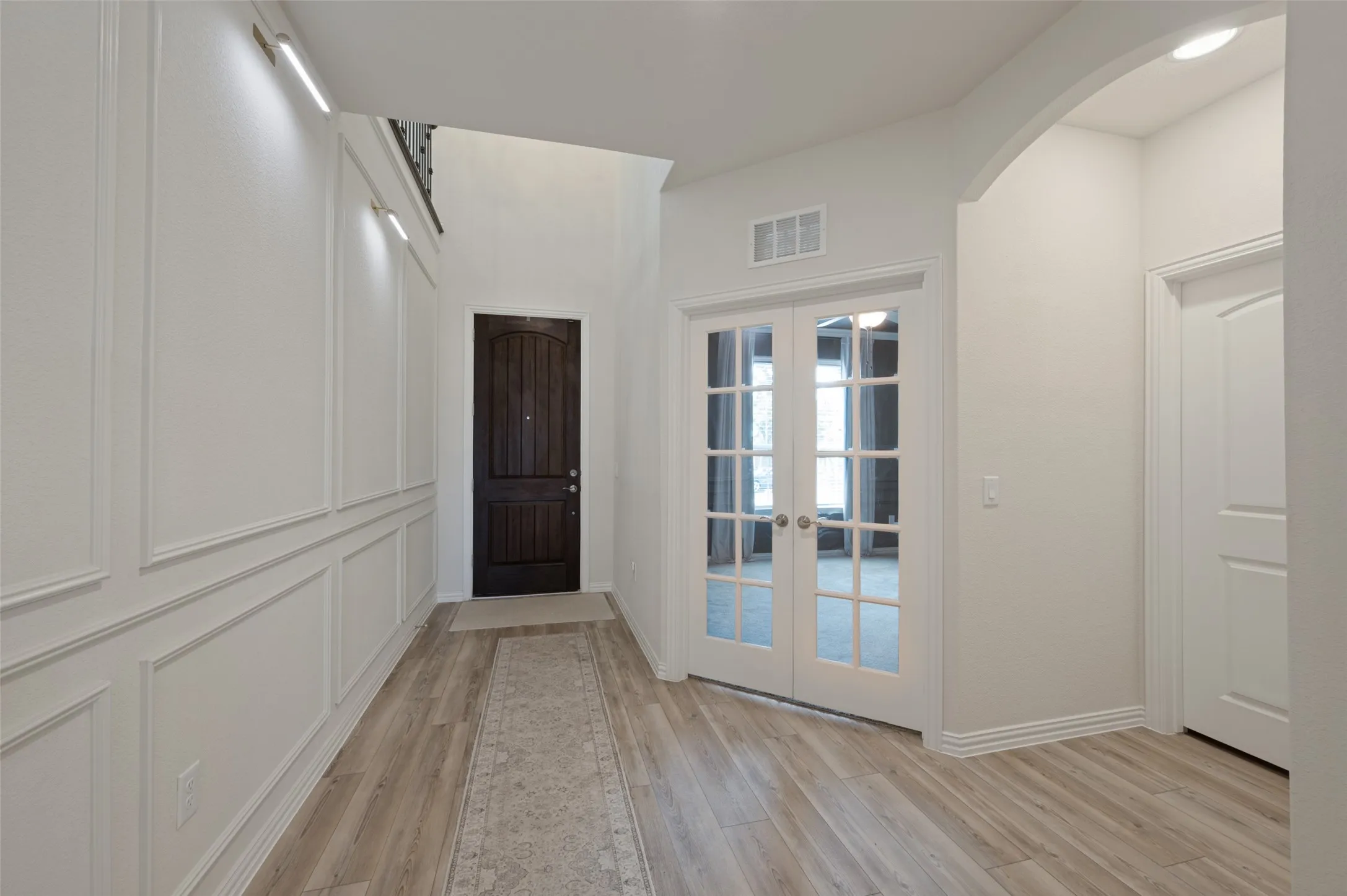 Wood-finish flooring, accent wall with decorative molding, dark wood entry door, white French doors with glass panels, and an arched doorway