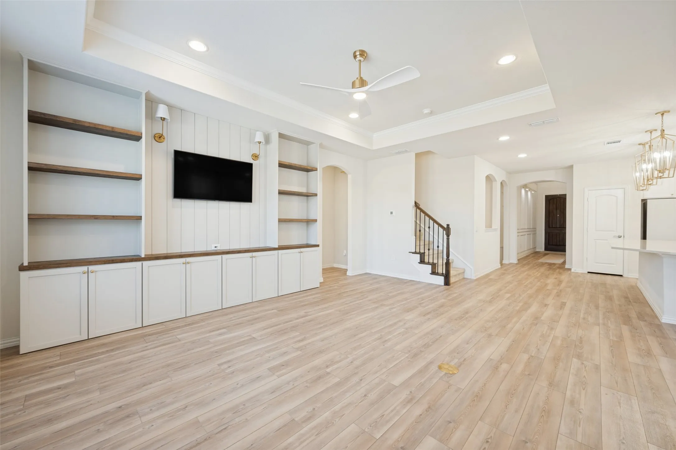Expansive living area with wood-finish flooring, built-in entertainment center with open shelving and cabinet storage, ceiling fan, recessed lighting, and crown molding