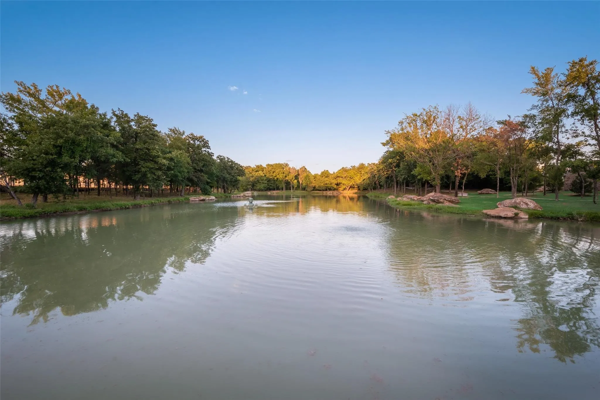 Expansive water feature with a central fountain, bordered by mature trees and natural rock landscaping on a manicured lawn