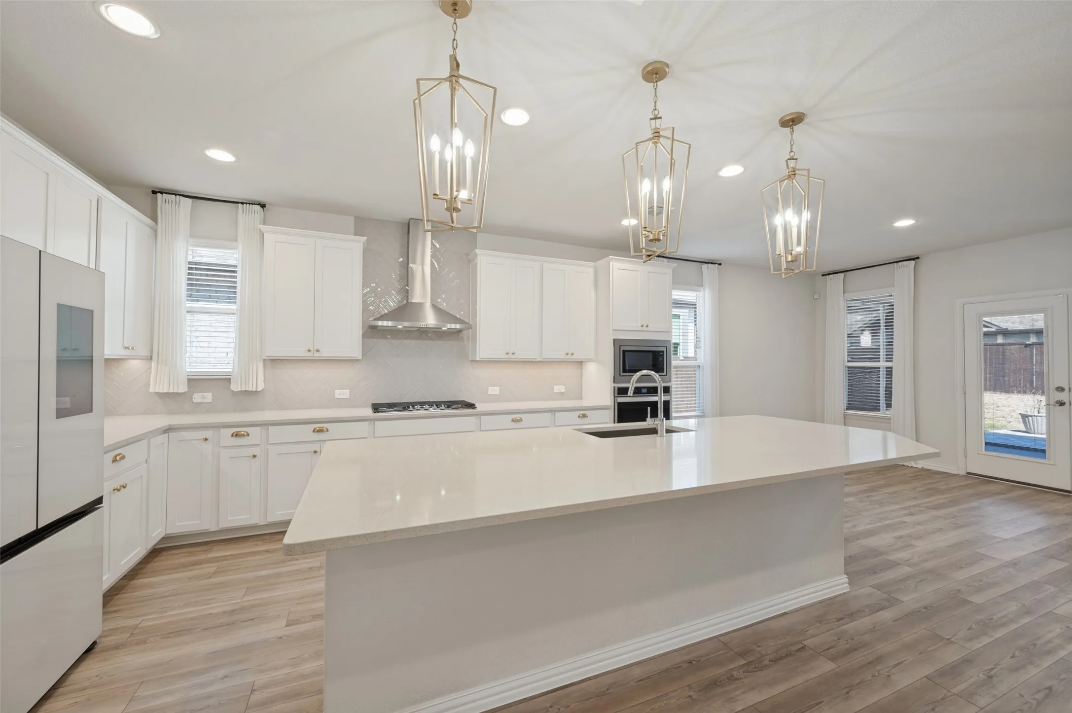 Kitchen featuring white cabinetry, light-toned countertops, a central island with sink, stainless steel appliances, and gold-finish pendant lighting