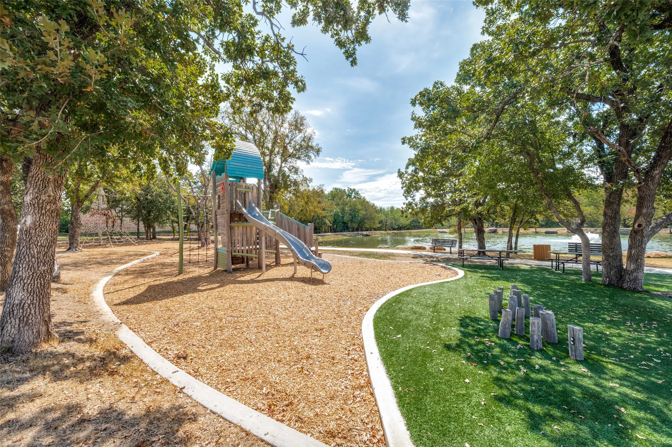 Playground area featuring a wood chip surface, artificial turf, and mature shade trees