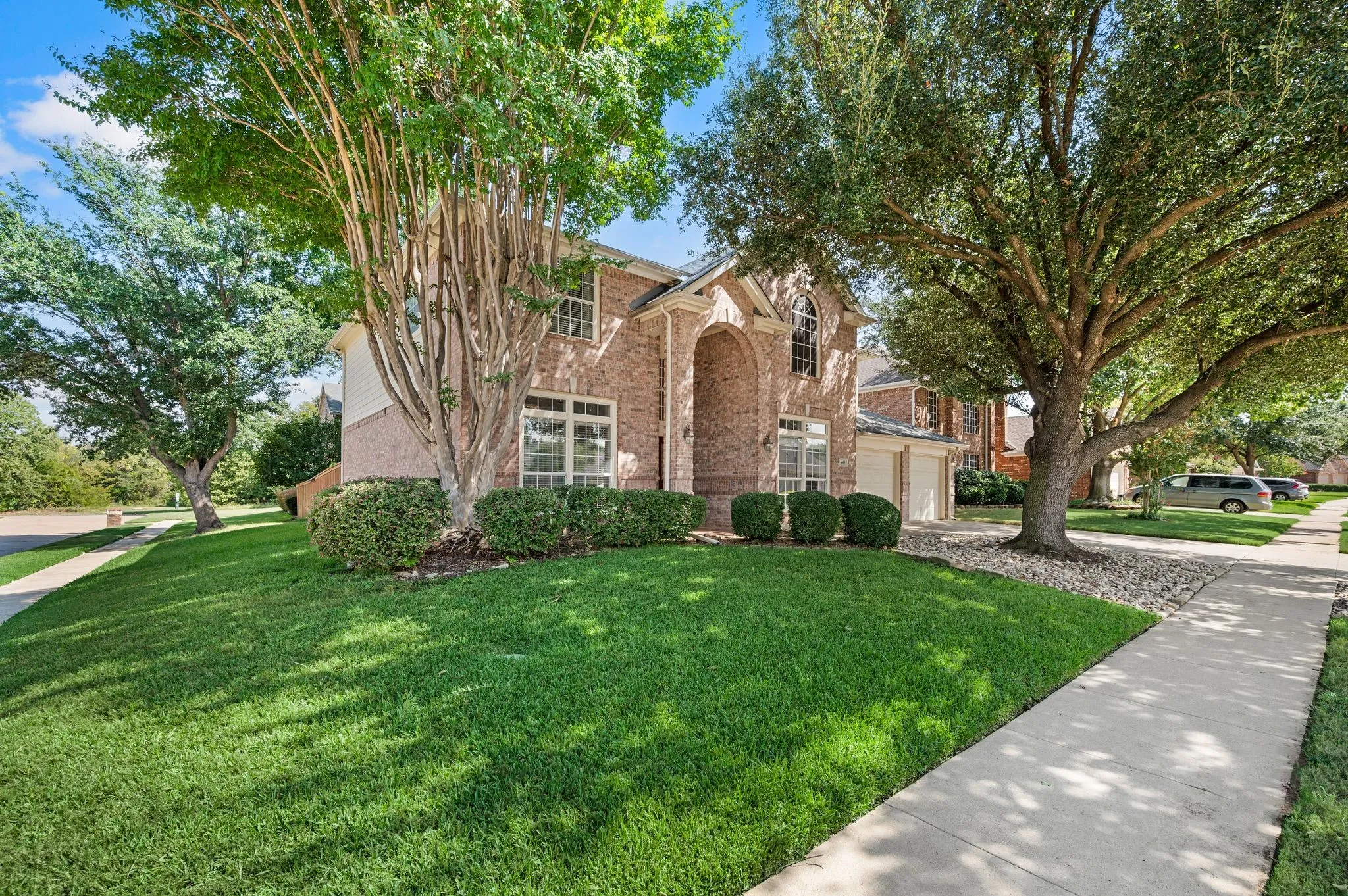 Brick exterior with arched entry, two-car garage, mature trees, and landscaped shrubbery