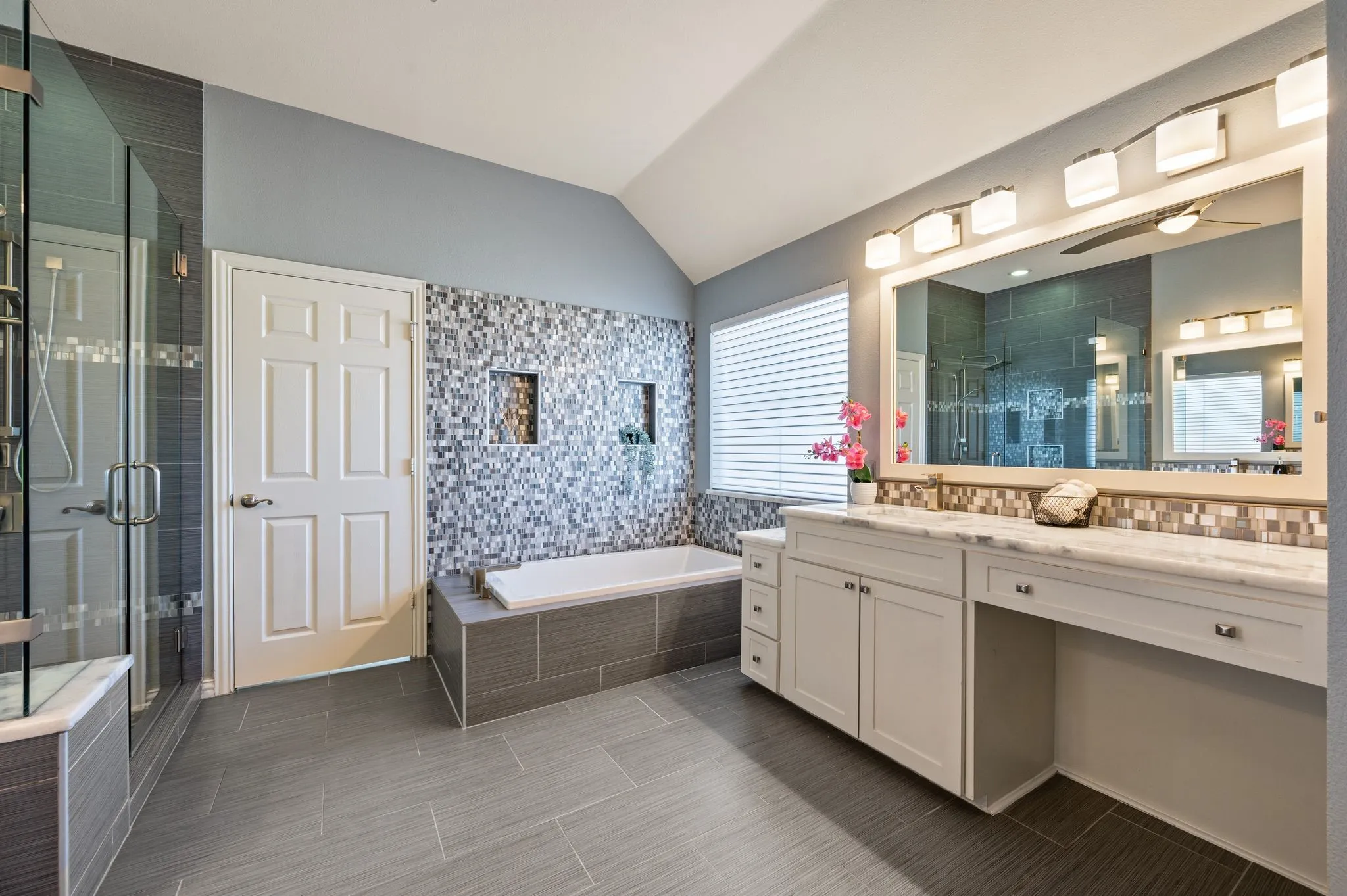 Vaulted ceiling bathroom featuring a glass-enclosed shower, built-in bathtub with mosaic tile surround, spacious vanity with double sinks, and integrated lighting