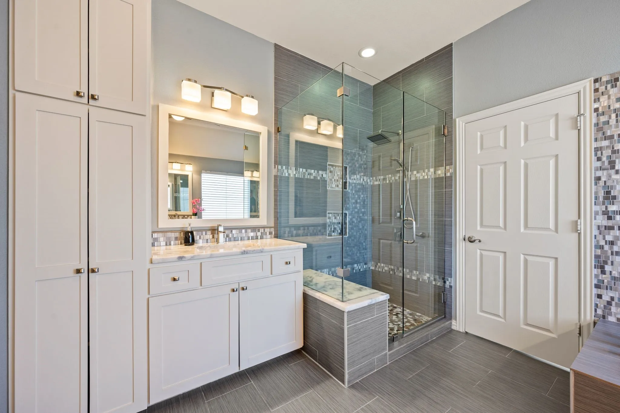 White shaker cabinetry, dual-sink vanity with marble countertop, glass-enclosed shower with bench, patterned tile accents, and large format floor tiling