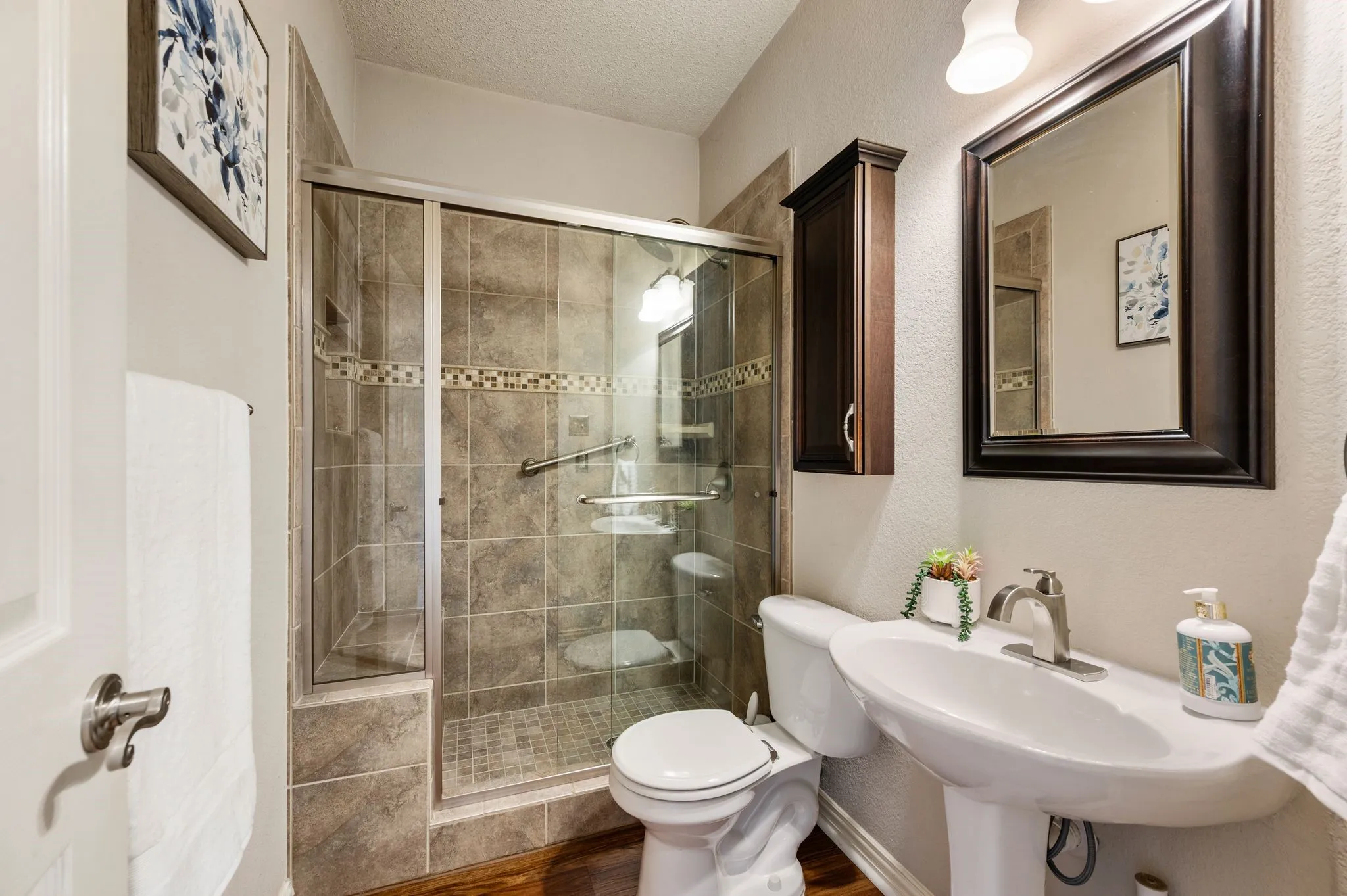 Pedestal sink, wood-finish flooring, walk-in shower with glass enclosure, wall-mounted storage cabinet, and framed mirror
