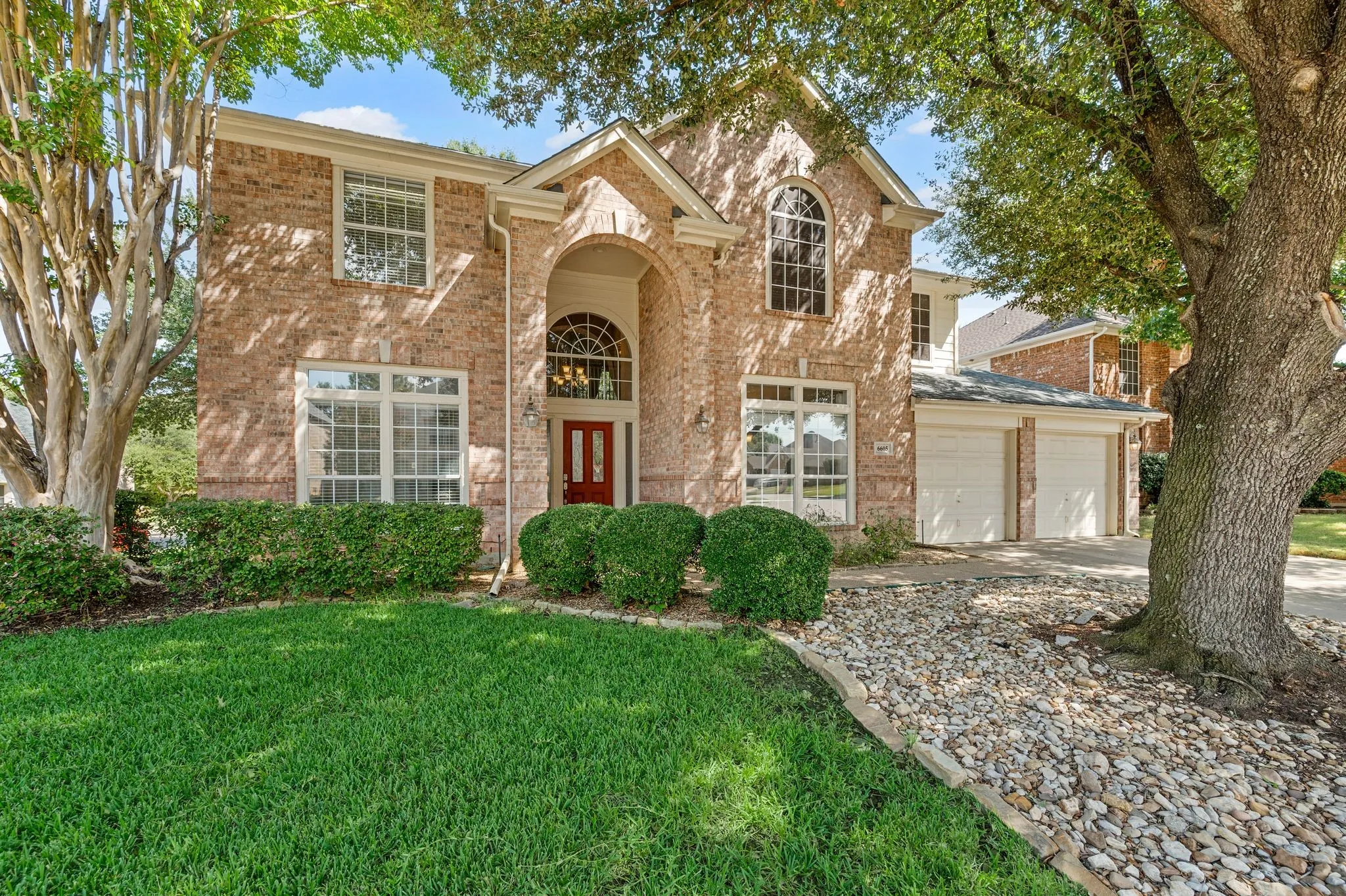 Brick exterior with arched entry, two-car garage, manicured lawn, and mature trees