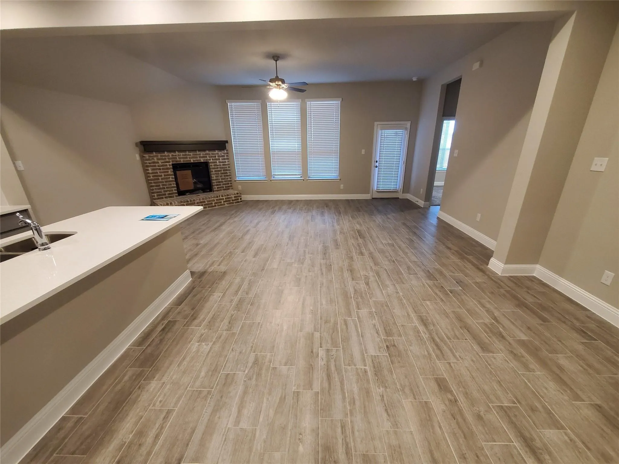 Spacious living area featuring wood-finish tile flooring, a brick fireplace with a wood mantle, and multiple windows with blinds