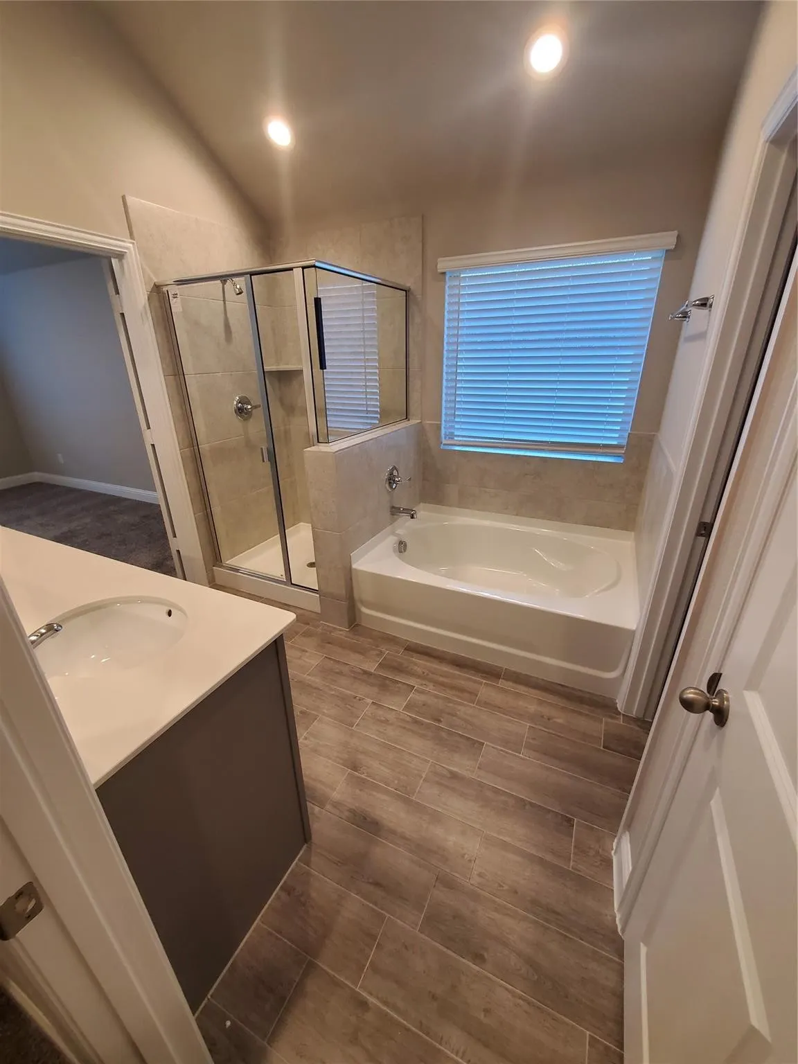 Walk-in shower with clear glass enclosure, built-in soaking tub, single vanity with integrated sink, light-colored countertop, and wood-finish tile flooring