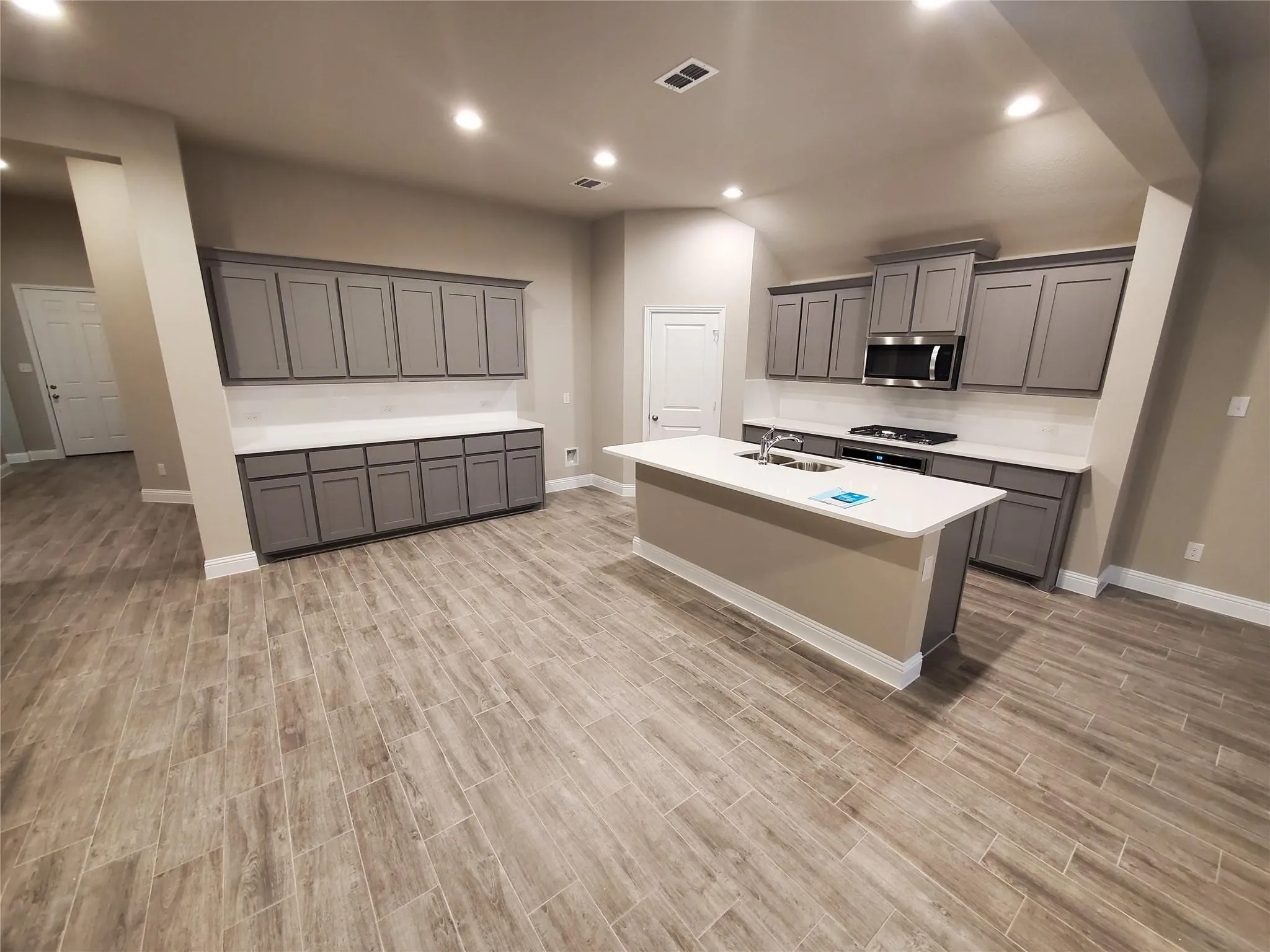 Open-concept kitchen featuring an island with a sink, stainless steel microwave, gray cabinetry, white countertops, and wood-finish tile flooring