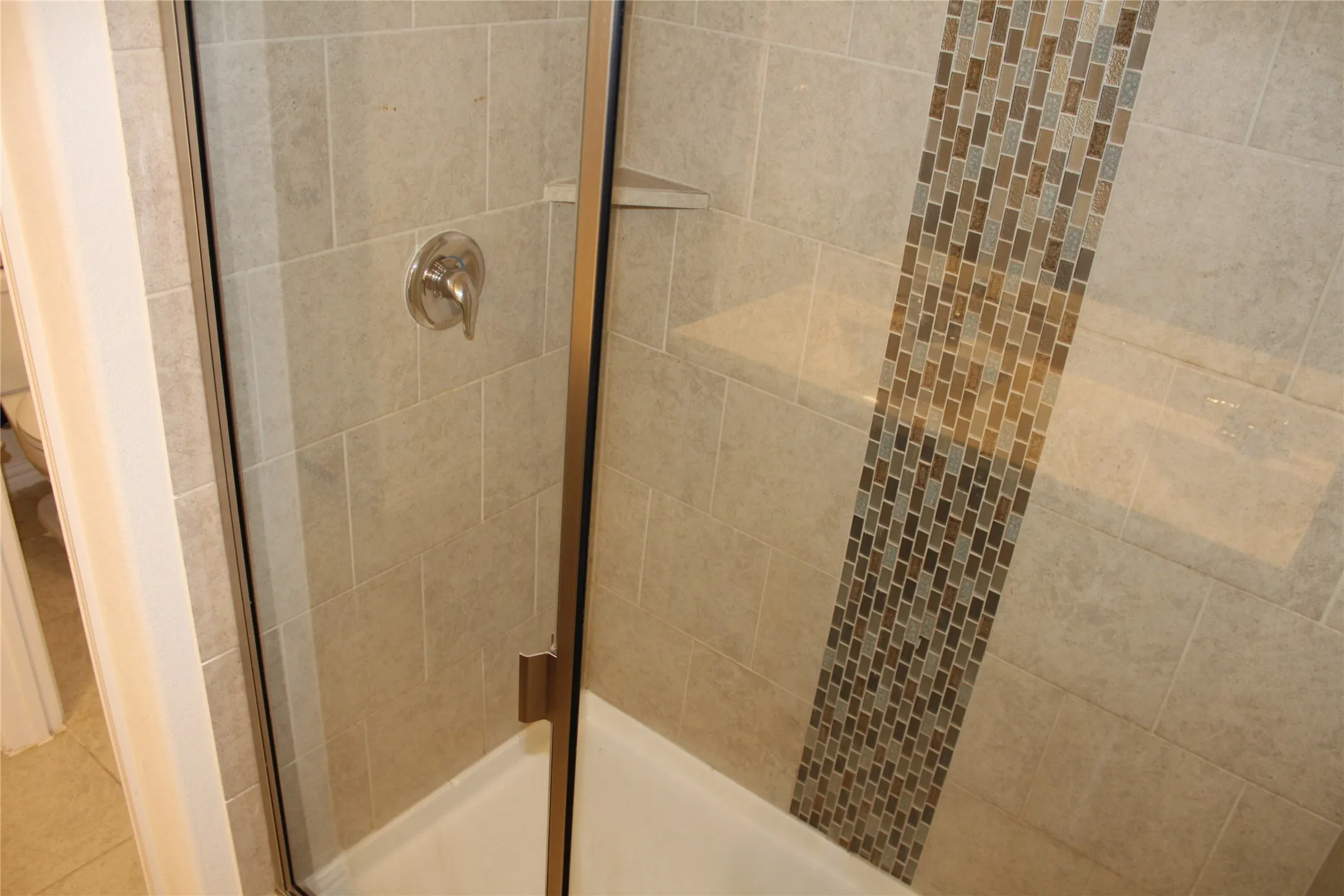 Tiled shower enclosure featuring a glass door, brushed nickel fixtures, an integrated corner shelf, and a decorative mosaic tile accent
