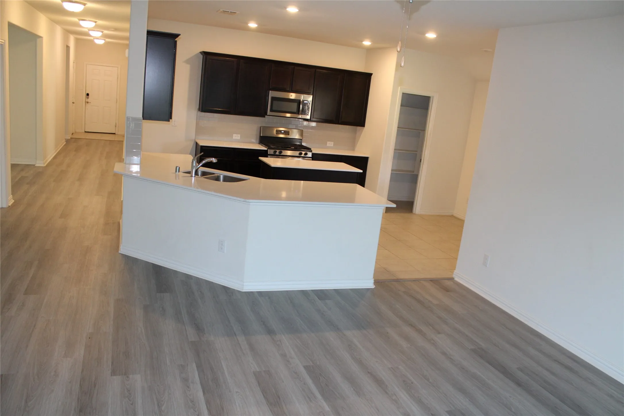 Kitchen featuring dark wood-finish cabinetry, stainless steel appliances, a white tile backsplash, and a central island with an integrated sink and light-toned countertop