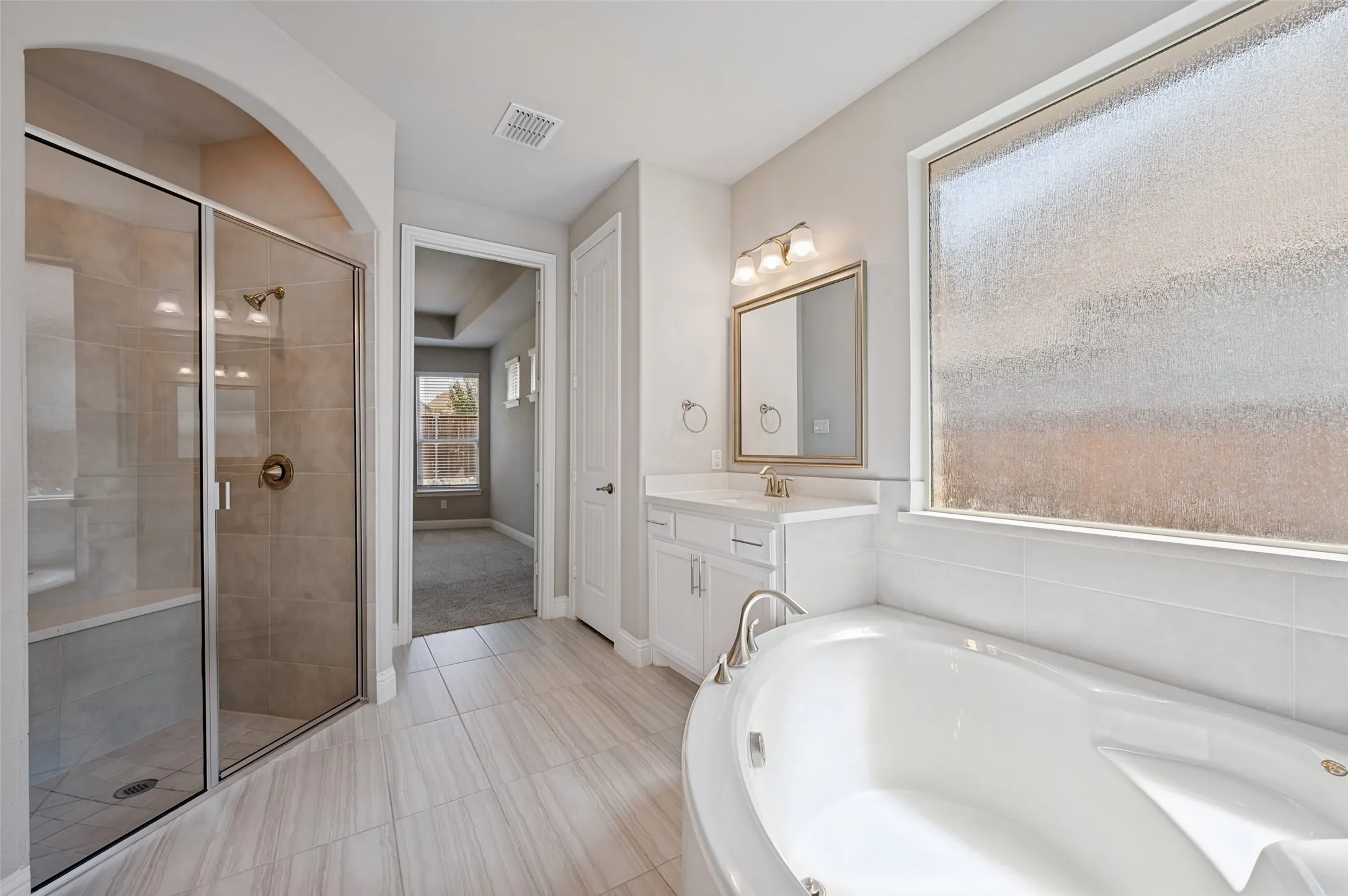 Curved soaking tub with adjacent window, separate glass-enclosed shower with tiled walls, wood-finish flooring, and a white vanity with an undermount sink