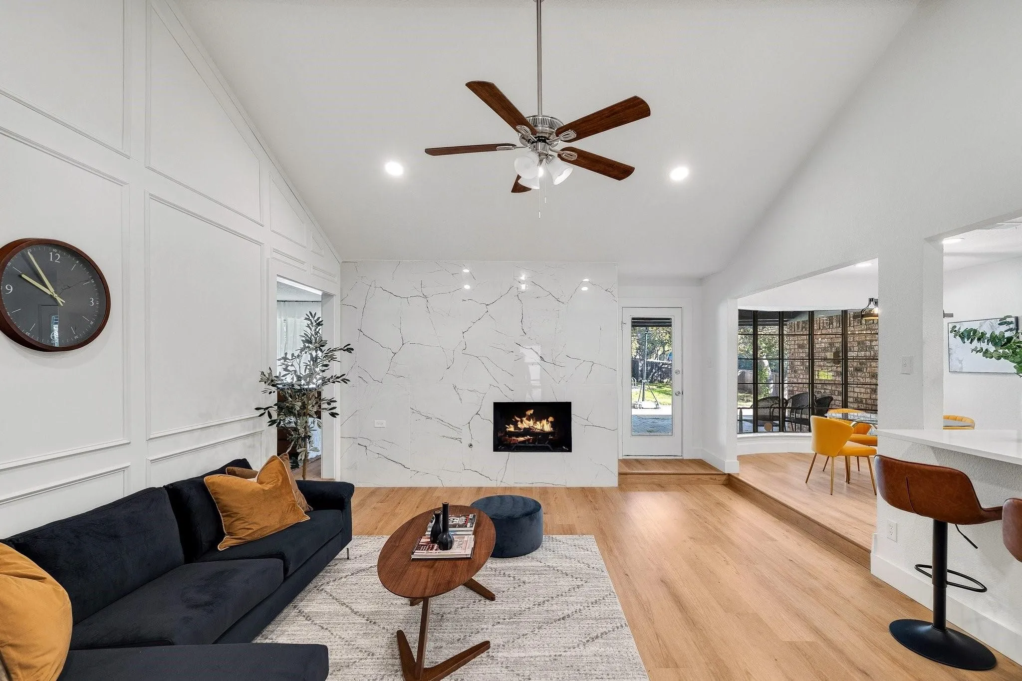 Vaulted ceiling with recessed lighting and ceiling fan, wood-finish flooring, accent wall with decorative molding, fireplace with stone-finish surround, and a raised dining area