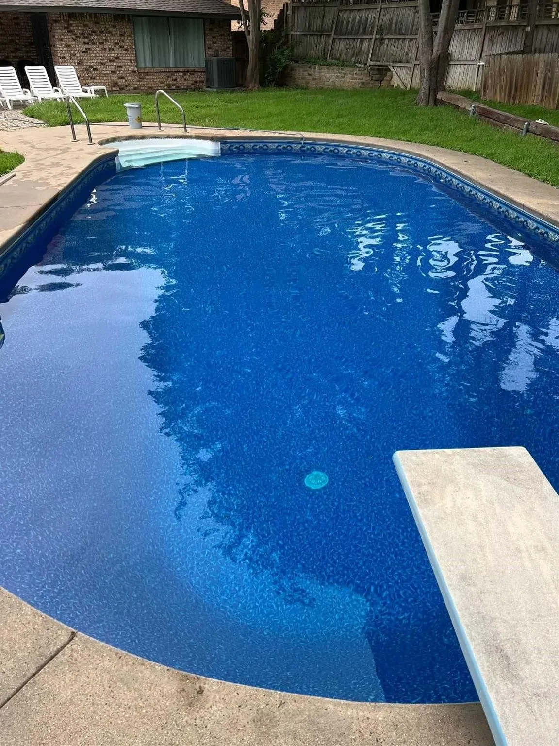 Oval-shaped swimming pool with a diving board, surrounding concrete decking, and steps with handrails