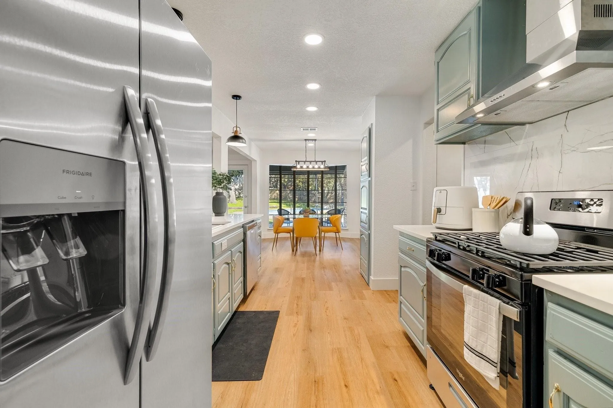 Stainless steel Frigidaire refrigerator, gas range with stainless steel hood, light wood-finish flooring, light blue cabinetry, and white countertops with a light veined backsplash