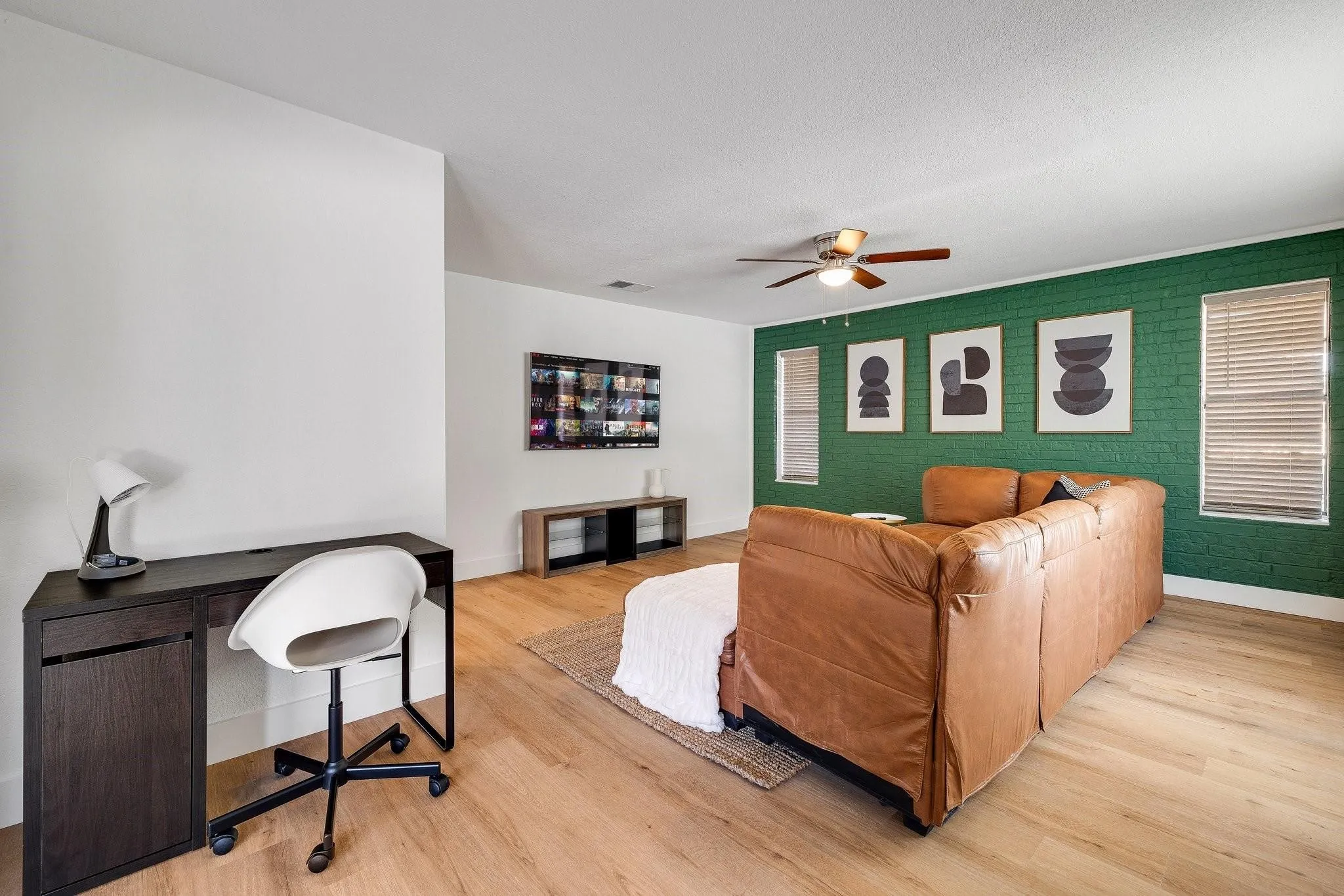 Light wood-finish flooring throughout, accent wall with painted brick texture, ceiling fan with integrated lighting, and multiple windows with blinds