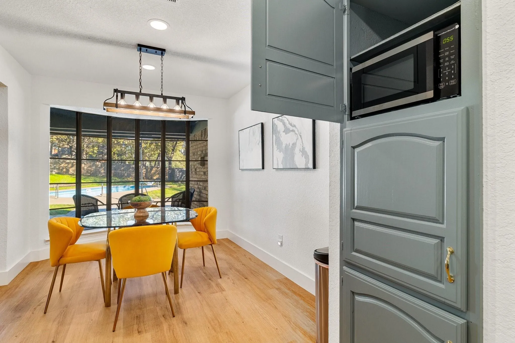 Hardwood-look flooring, bay window with exterior scenery, overhead multi-bulb linear light fixture, recessed lighting, and built-in cabinetry with integrated microwave
