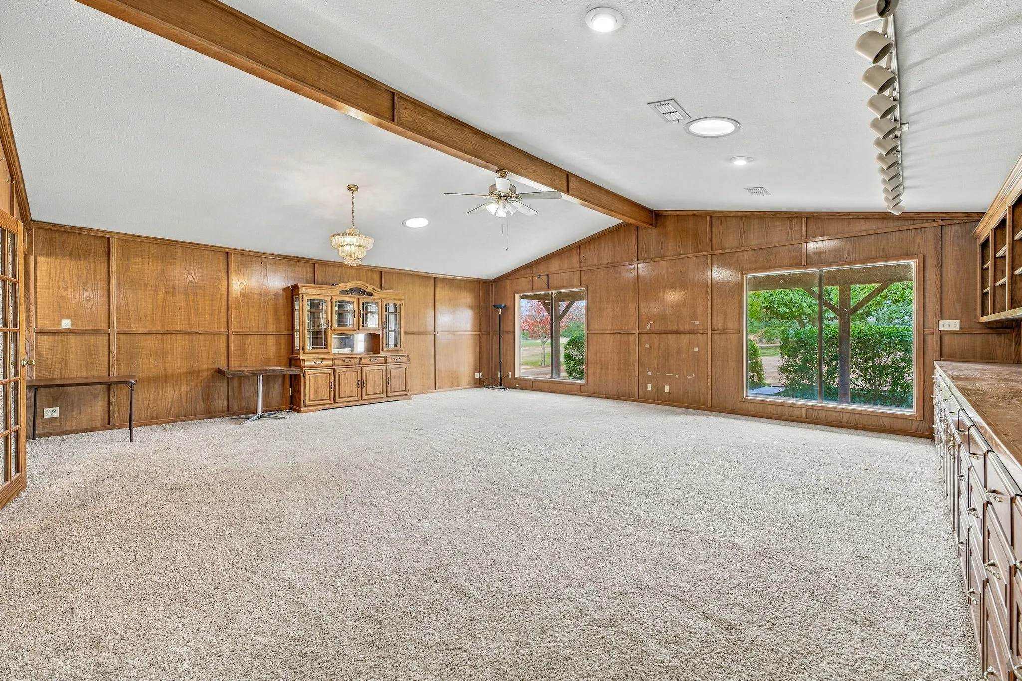 Spacious interior featuring wood-paneled walls, vaulted ceiling with exposed beam, expansive windows, recessed lighting, and a crystal chandelier