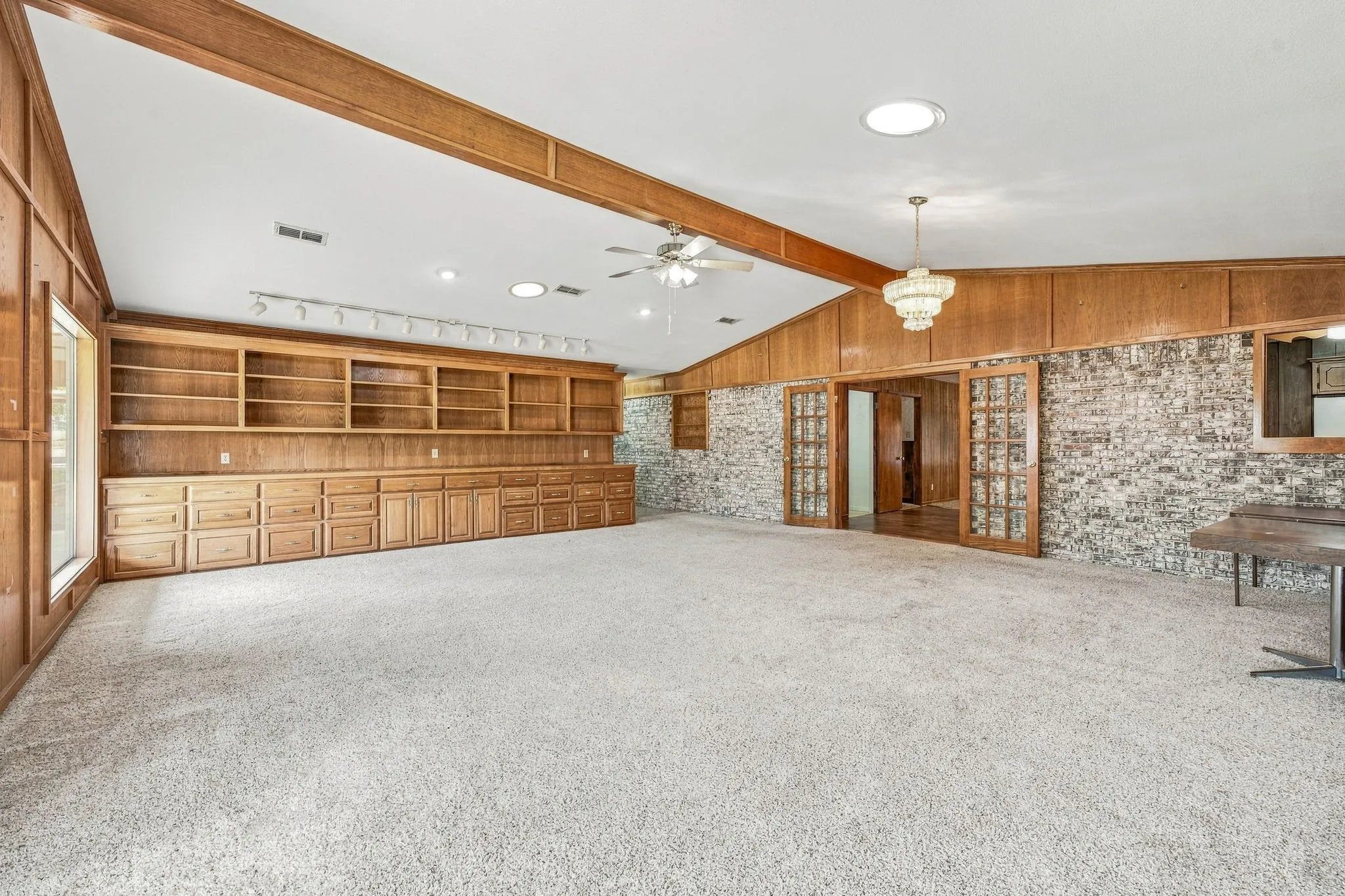 Vaulted ceiling with exposed wood beams, extensive built-in wooden cabinetry and shelving, brick accent wall, double glass-paneled doors, and recessed​​‌​​​​‌​​‌‌​‌‌​​​‌‌​‌​‌​‌​​​‌​​ lighting
