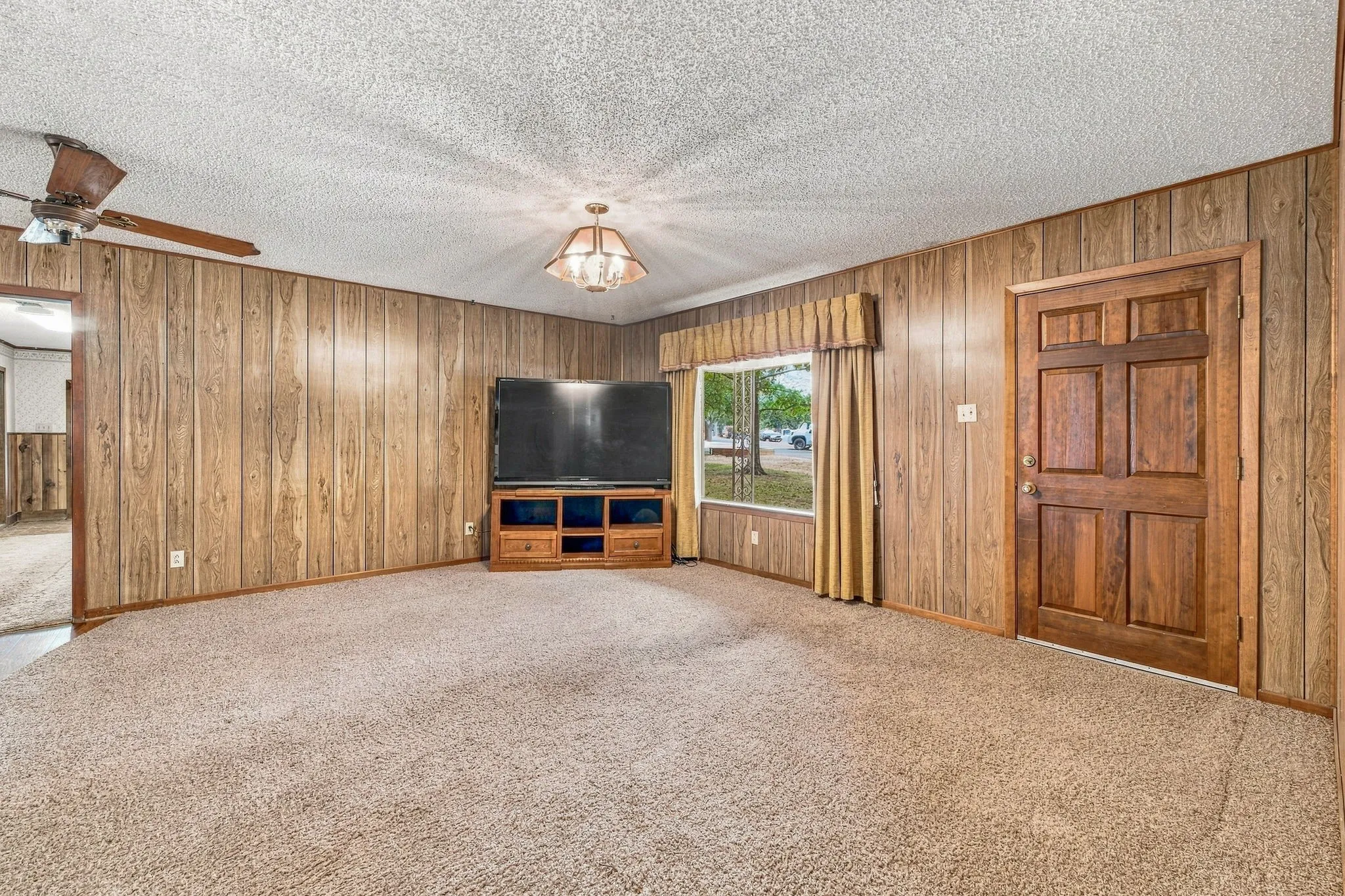 Carpeted living area featuring wood-paneled walls, an overhead light fixture, a ceiling fan, and a paneled entry door