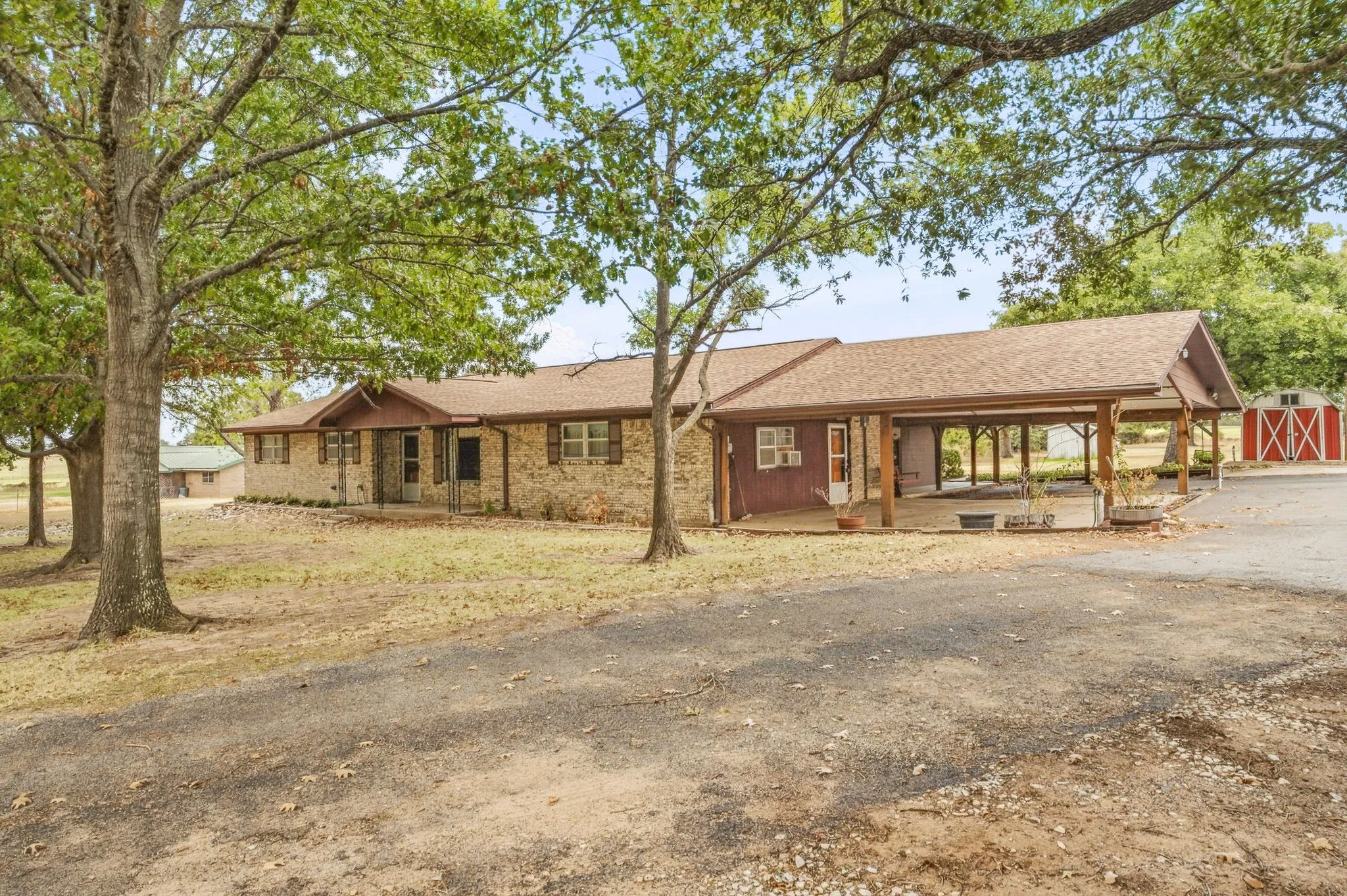 Brick exterior, covered porch entry, carport structure, mature trees, and gravel driveway