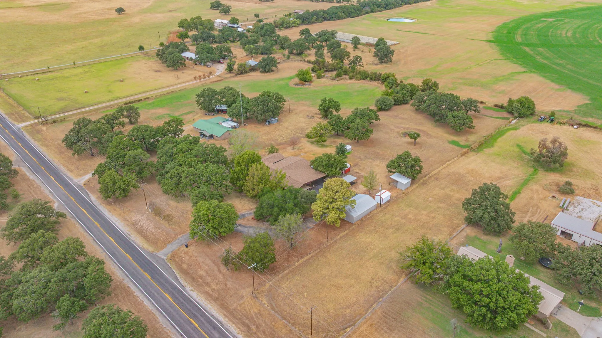Expansive rural landscape with a paved road, scattered mature trees, and a visible circular irrigation pattern in a distant field