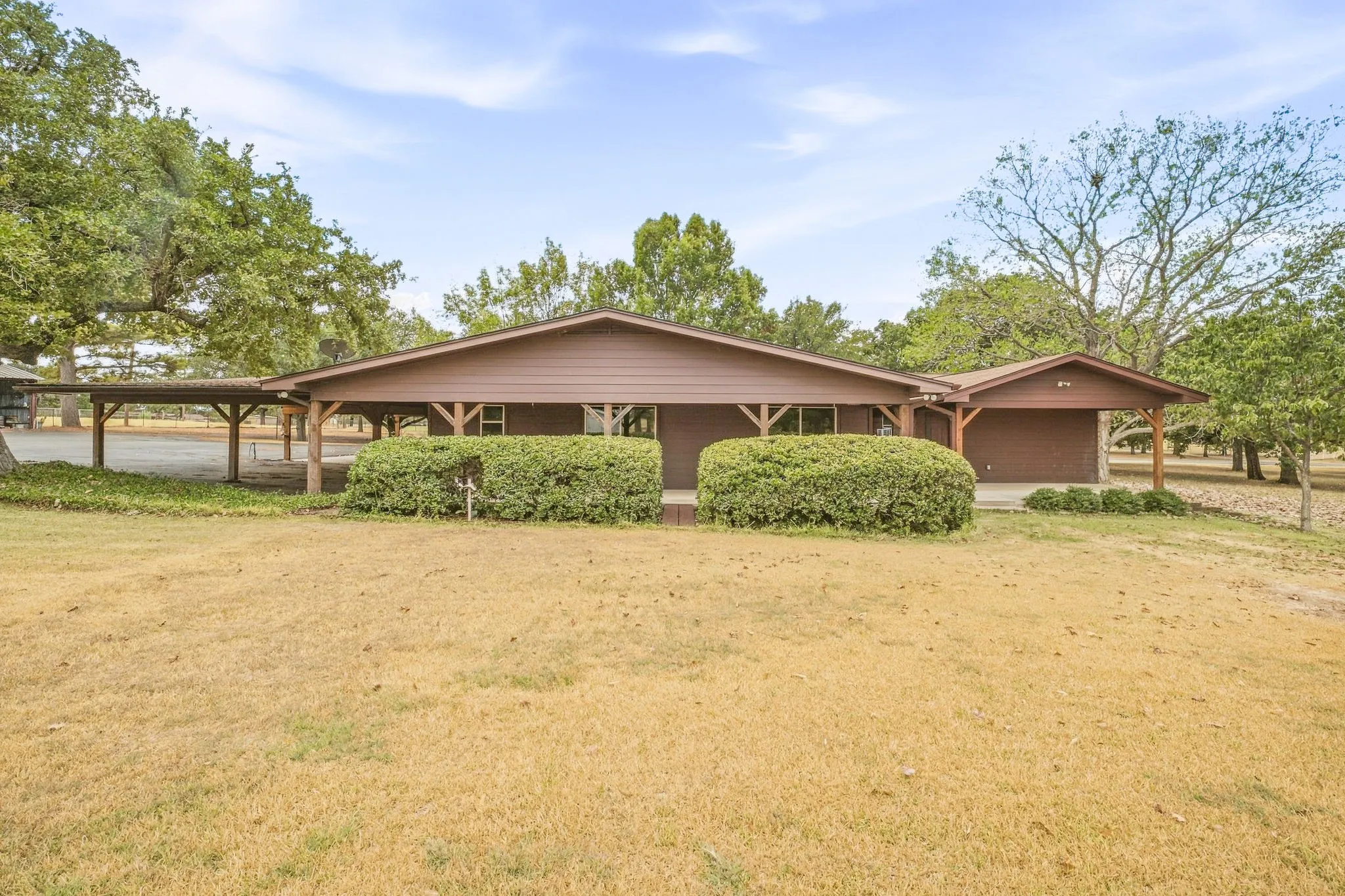 Brown siding exterior, covered front porch, integrated carport, mature landscaping, and an expansive lawn