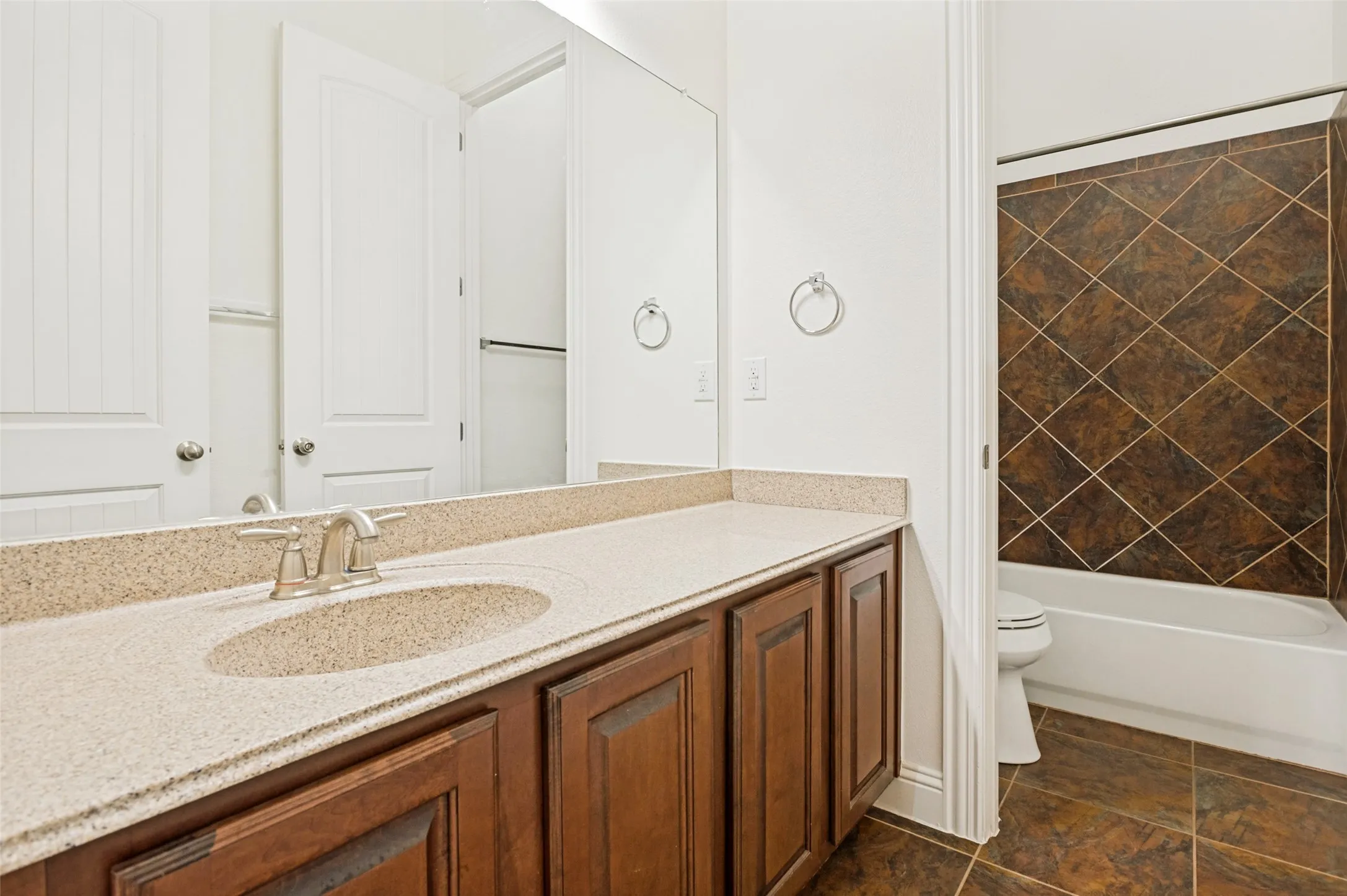 Bathroom featuring a light-colored countertop with an integrated sink, dark wood-finish cabinetry, a brushed nickel faucet, a full-width mirror, and a shower-tub combination with diagonal brown tile surround