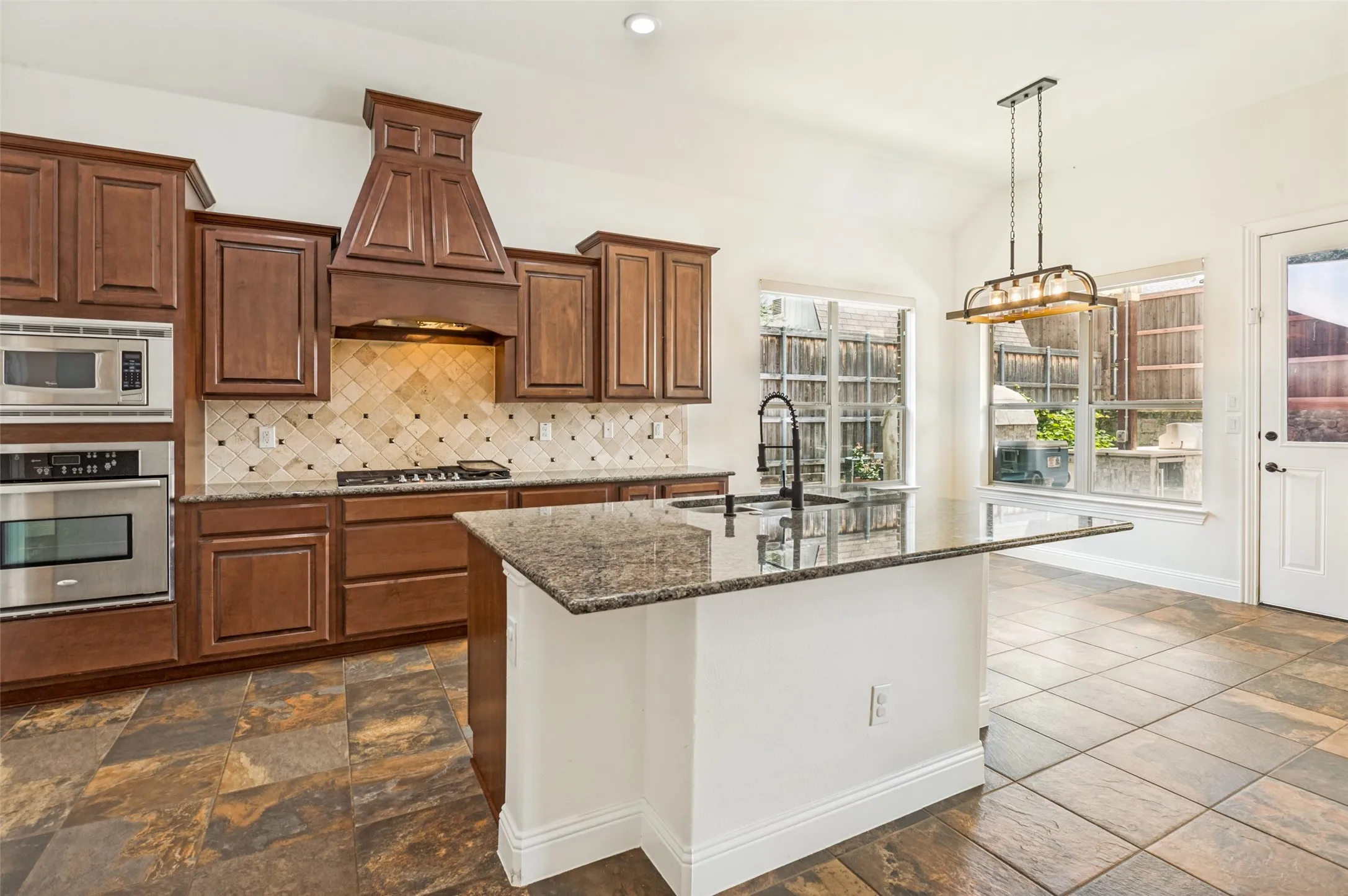Kitchen featuring wood cabinetry, stainless steel appliances, a tile backsplash, and a central island with a sink and granite countertop