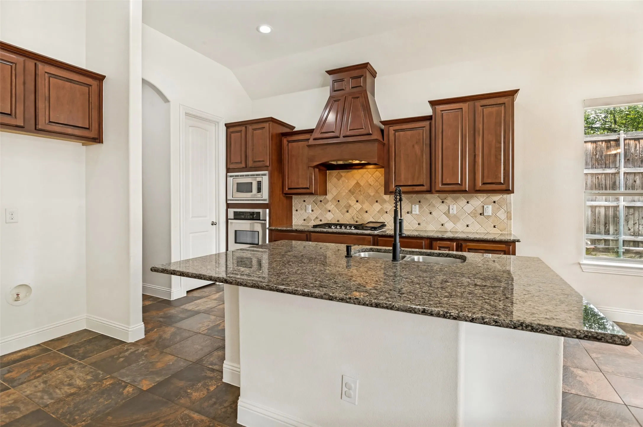 Kitchen featuring a granite-topped island with an integrated sink, rich wood cabinetry, stainless steel double wall ovens, a tiled backsplash, and stone-look flooring