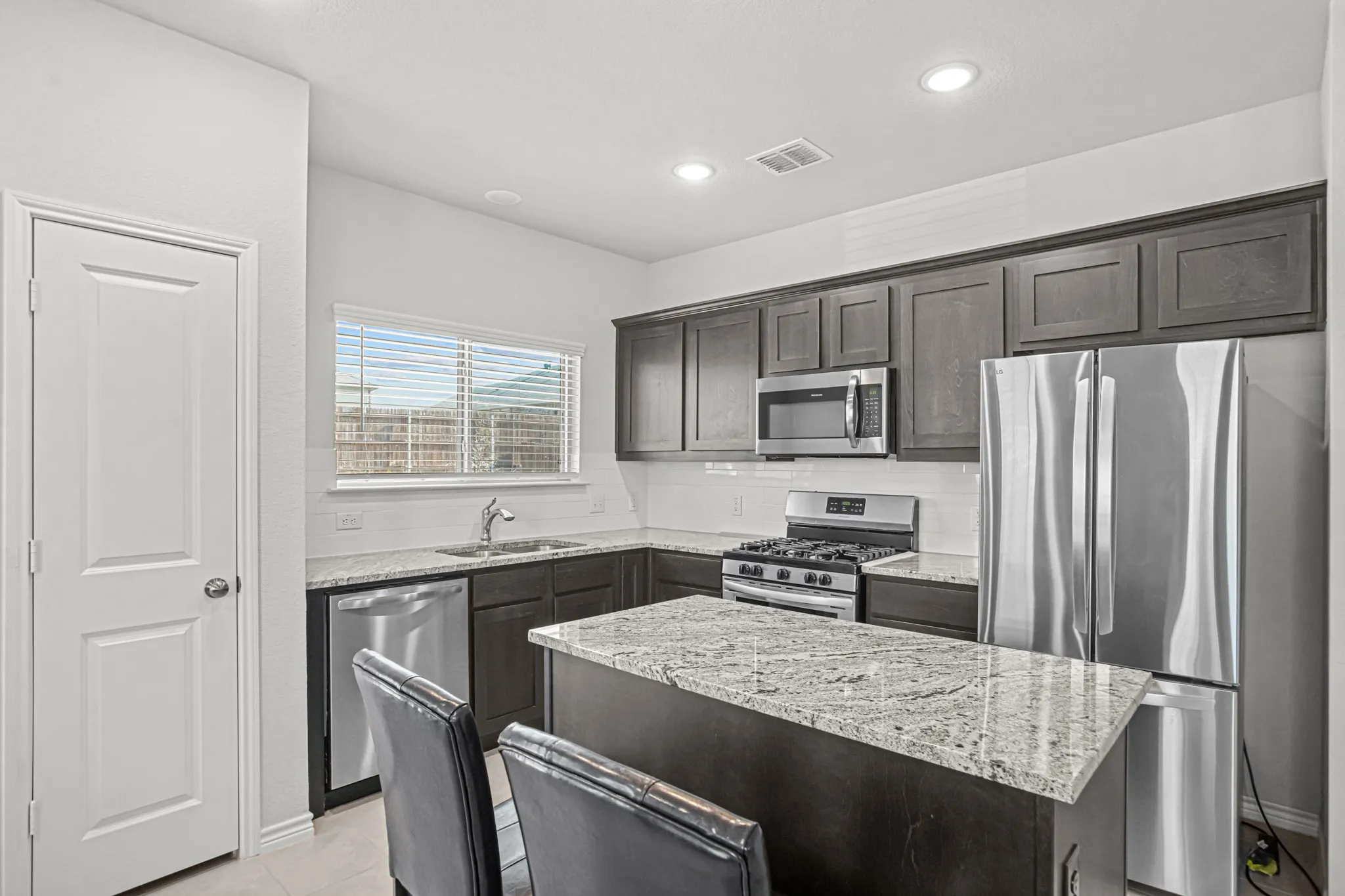 Kitchen featuring dark wood cabinetry, stainless steel appliances, light granite countertops, white subway tile backsplash, and recessed lighting