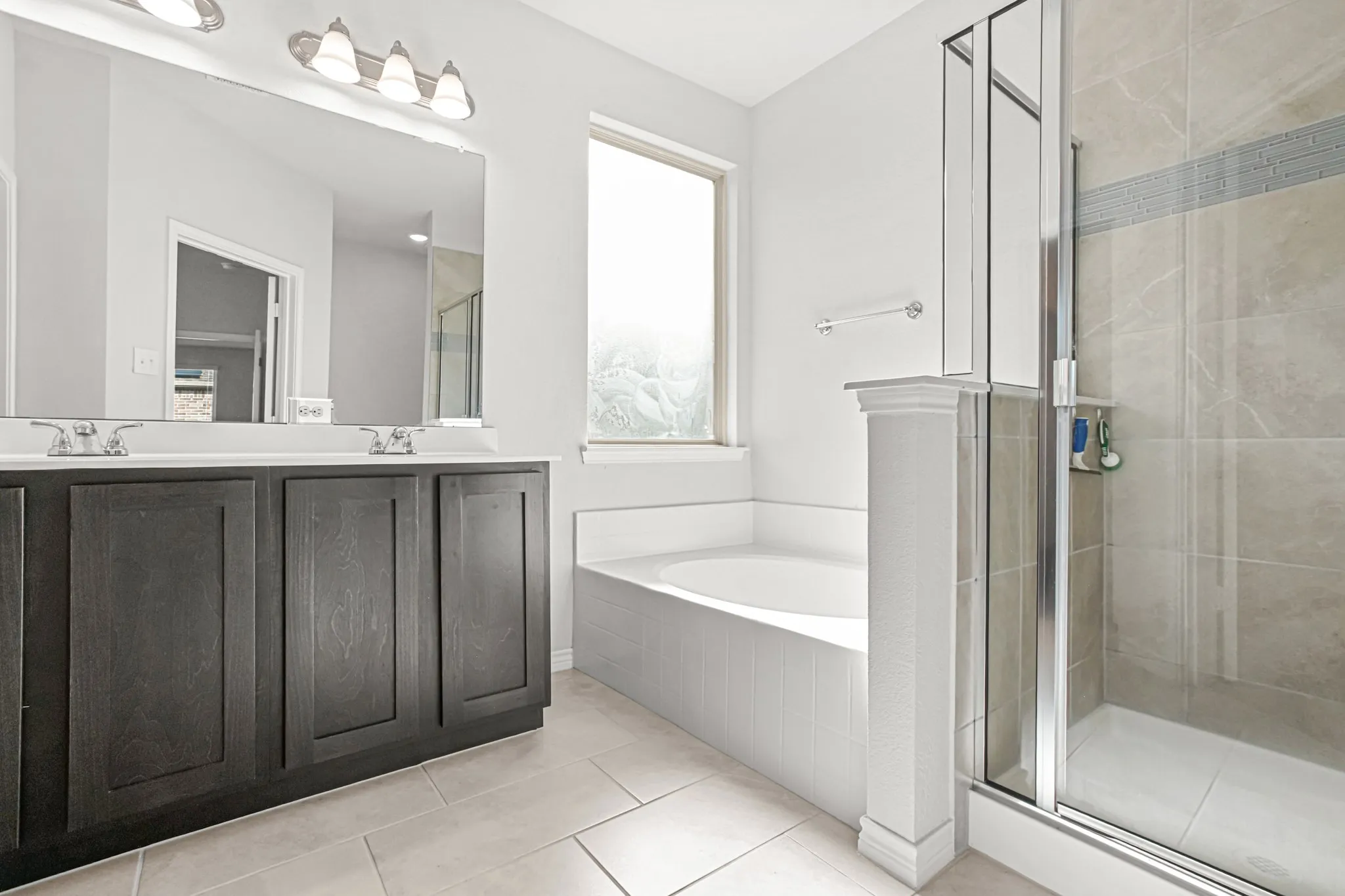 Double vanity with dark wood-finish cabinetry, built-in soaking tub, walk-in shower with clear glass enclosure, and large window