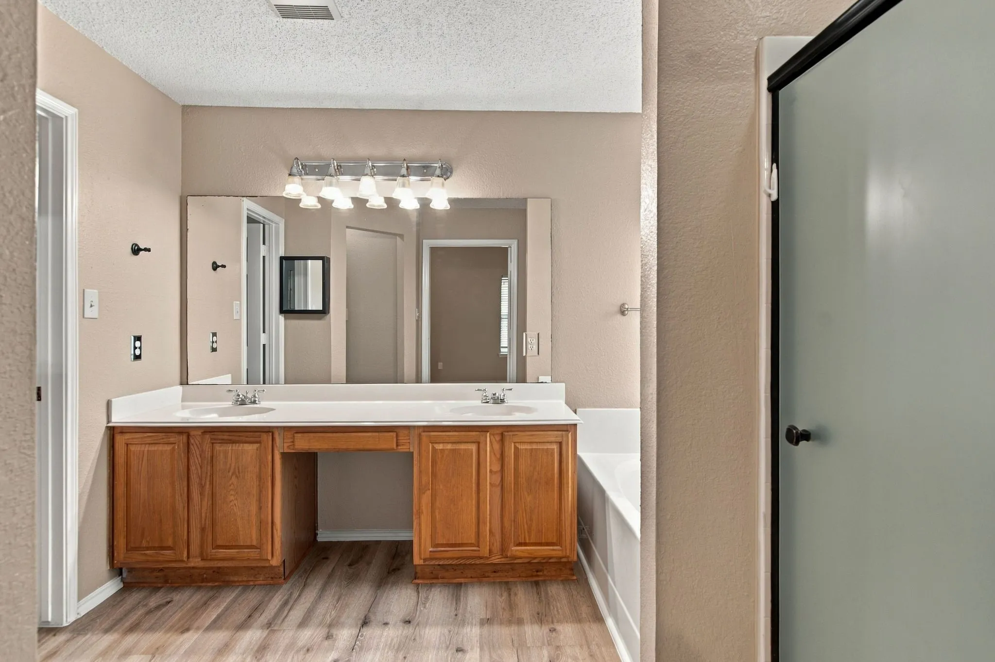 Wood-finish flooring, dual vanity with wood cabinetry, white countertops, built-in bathtub, and a frosted glass shower enclosure