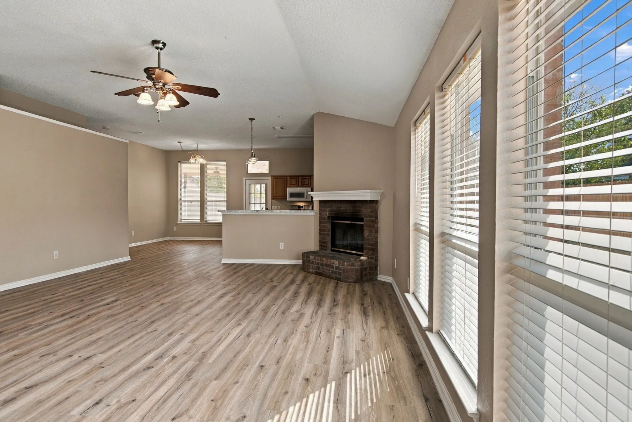Vaulted ceiling, ceiling fan, wood-finish flooring, brick fireplace, and multiple windows with blinds