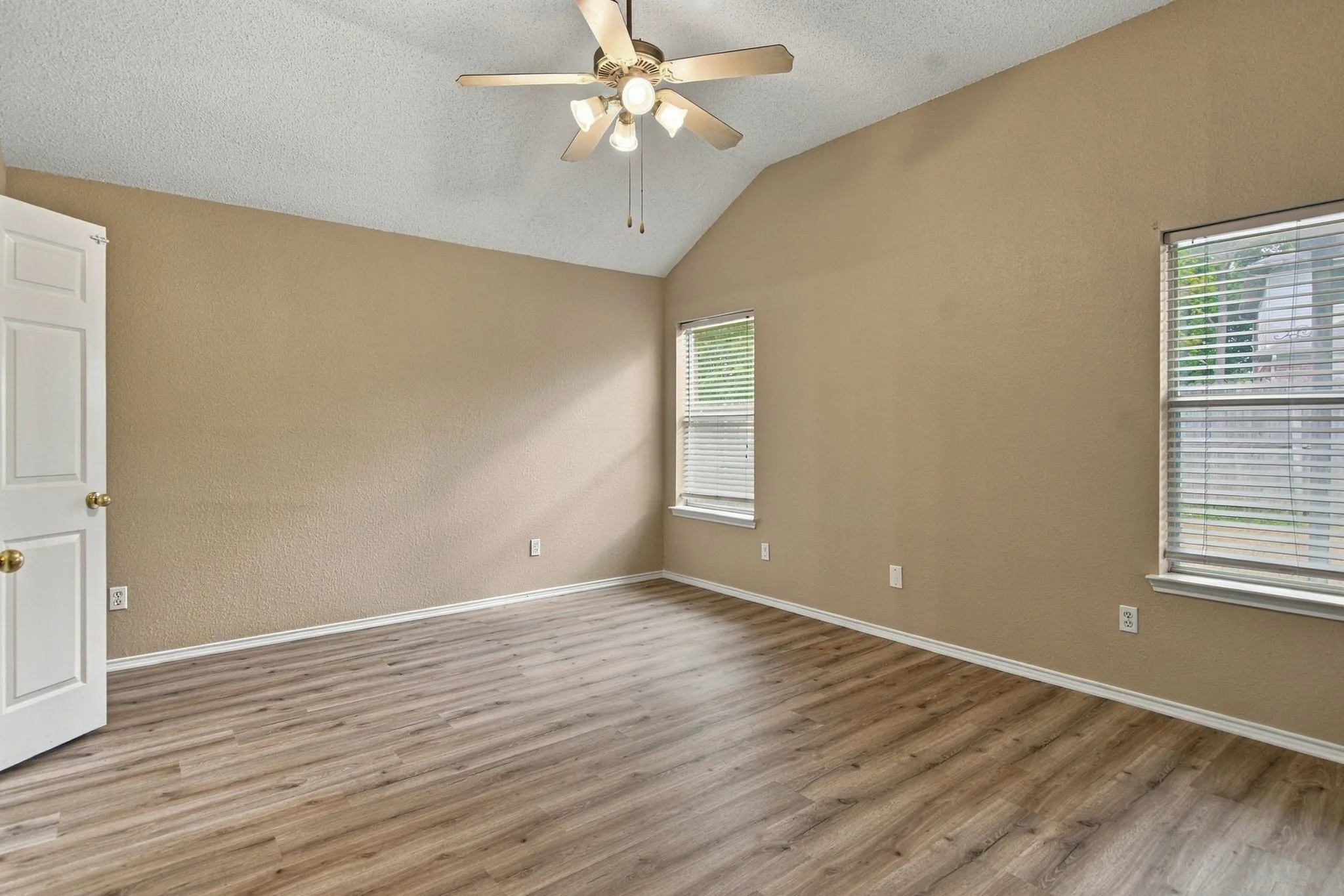 Vaulted ceiling, wood-finish flooring, two windows with blinds, ceiling fan with integrated lighting, and a paneled door